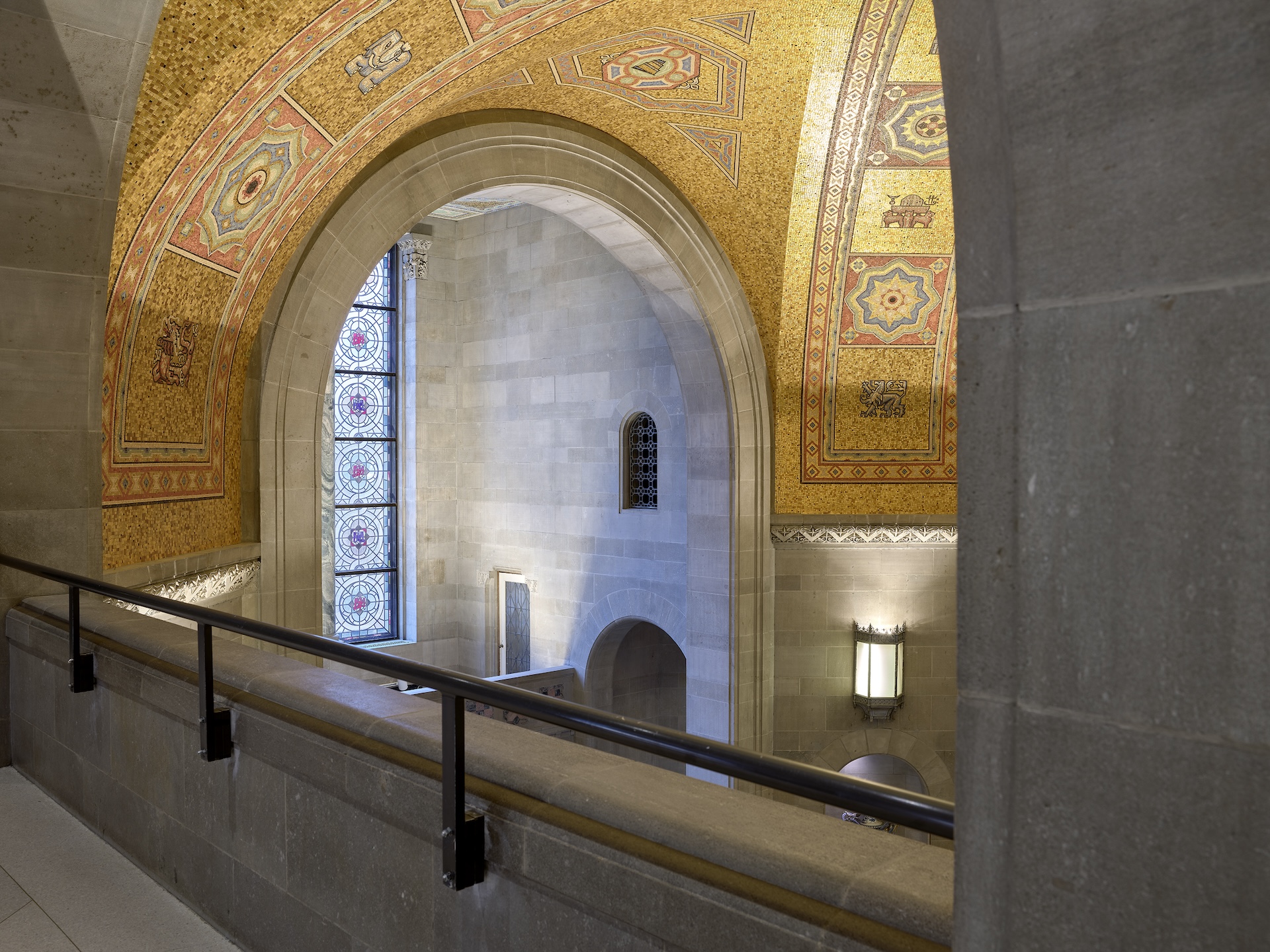 Stone interior arches and walls with a mosaic gold-patterned arch above and a metal railing in front.