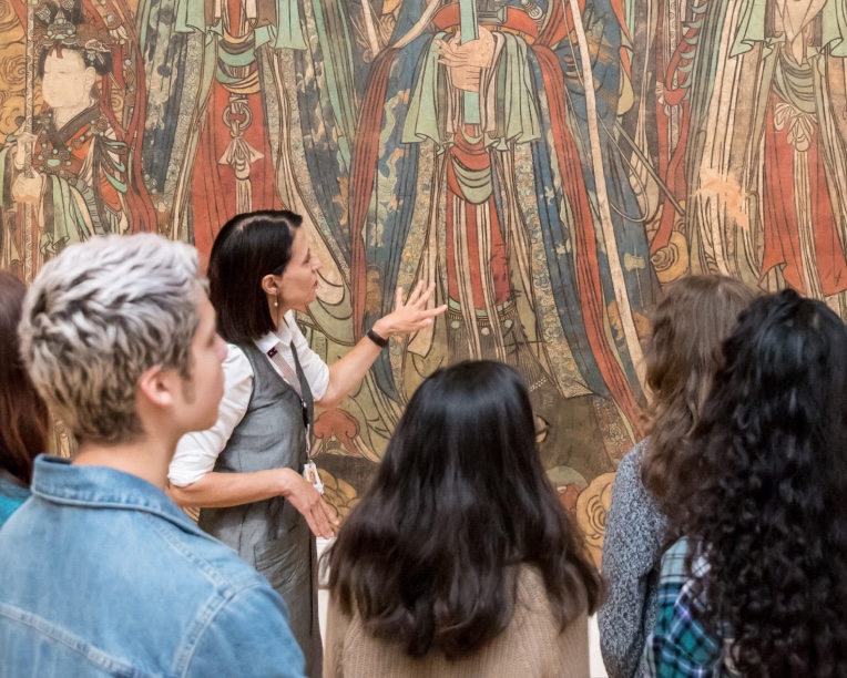 A group of four people view and discuss a large, colorful mural or painting featuring figures in traditional robes.