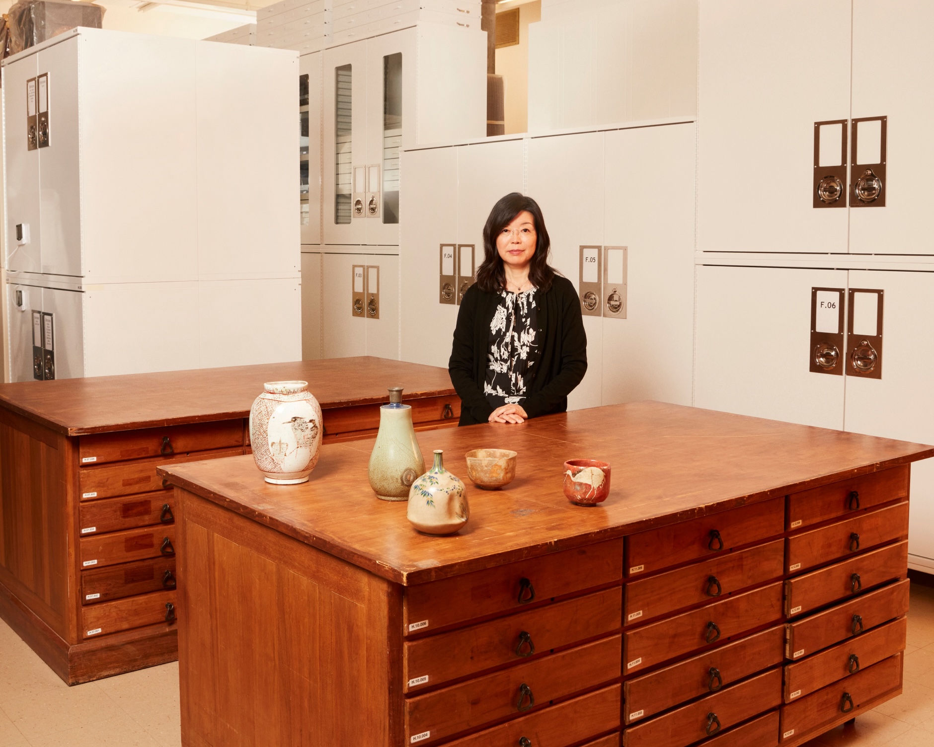 A woman stands behind a wooden cabinet with drawers, displaying five ceramic vessels of varying shapes and sizes.