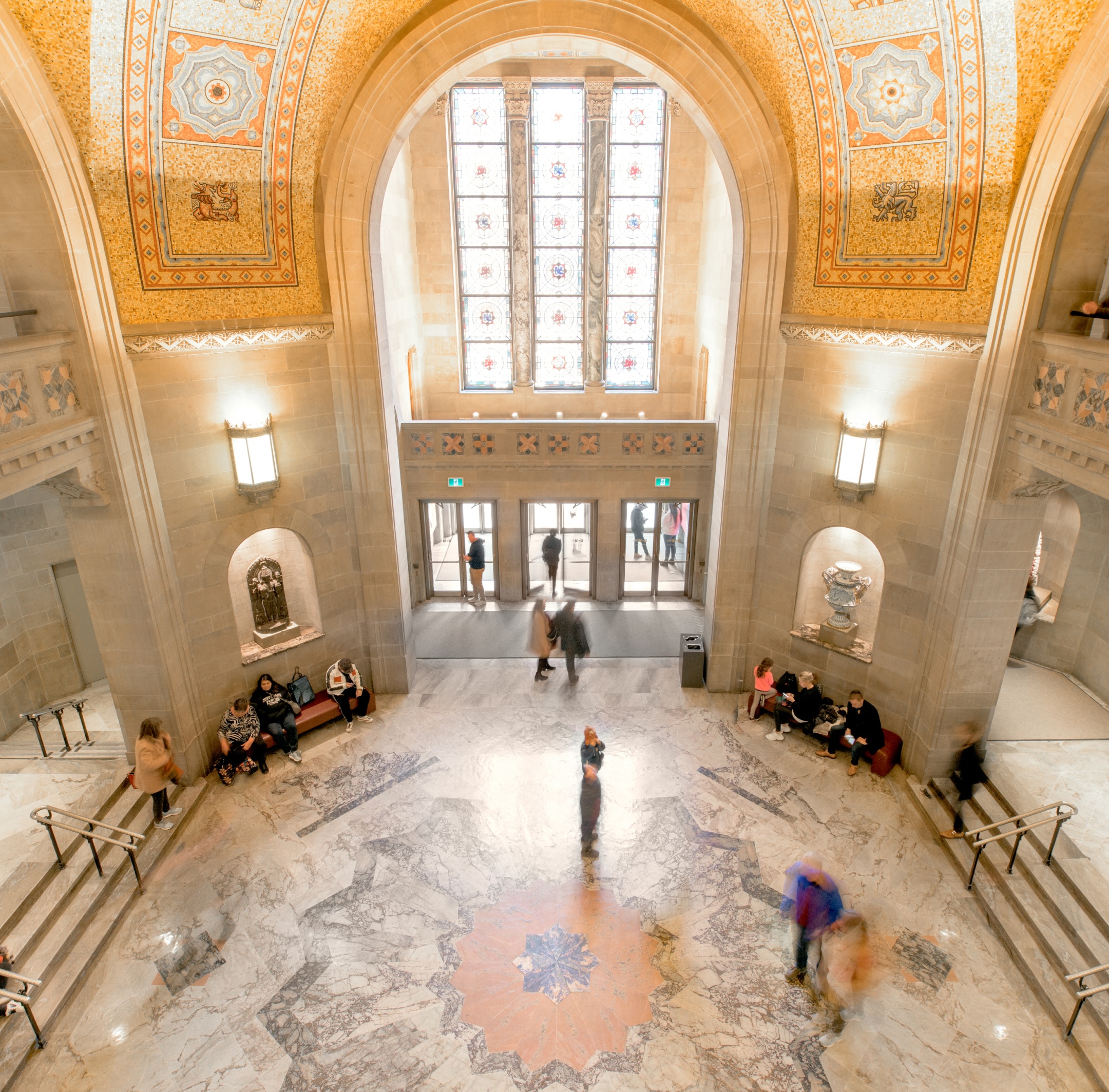 Spacious museum entrance hall with high arched ceiling, detailed wall carvings, and symmetrical seating areas along the sides.