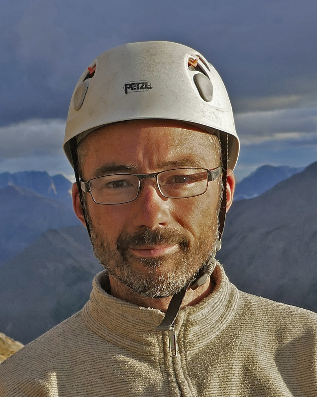 Man wearing glasses and a white Petzl helmet with a cloudy sky and mountains in the background.