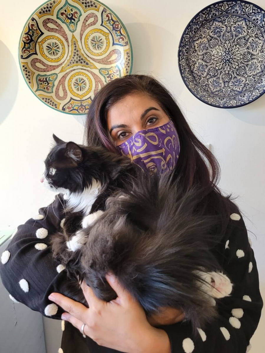 A woman wearing a purple patterned face mask and black polka-dot sweater holds two black and white cats in front of two decorative plates.