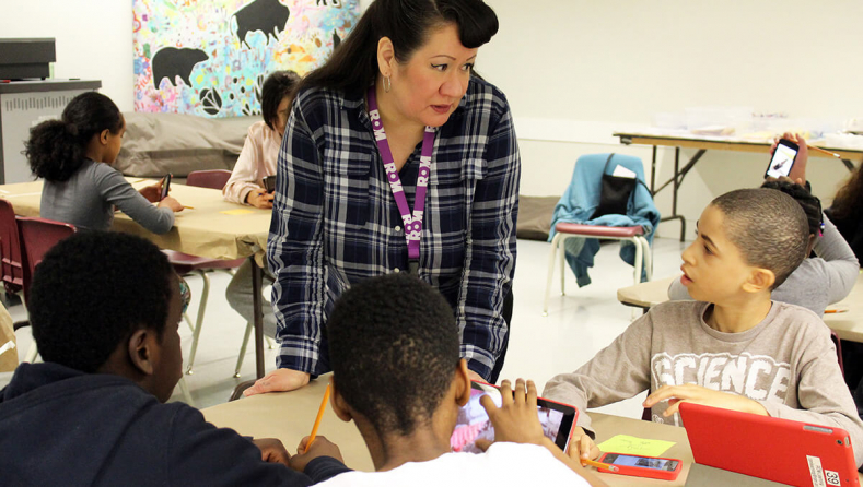 A teacher in a plaid shirt engages with four students seated around a table in a classroom.