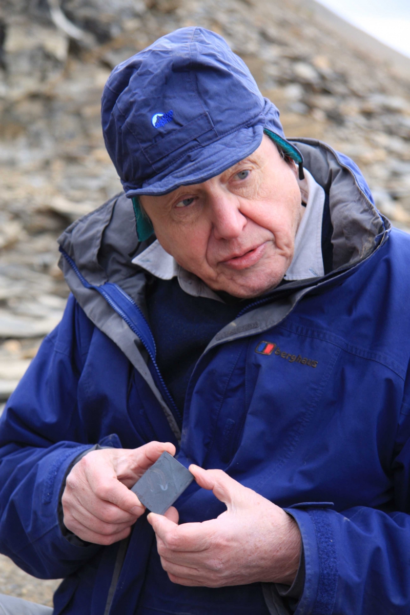 Man in a blue jacket and blue hat holding a small black object, outdoors near a rocky surface.