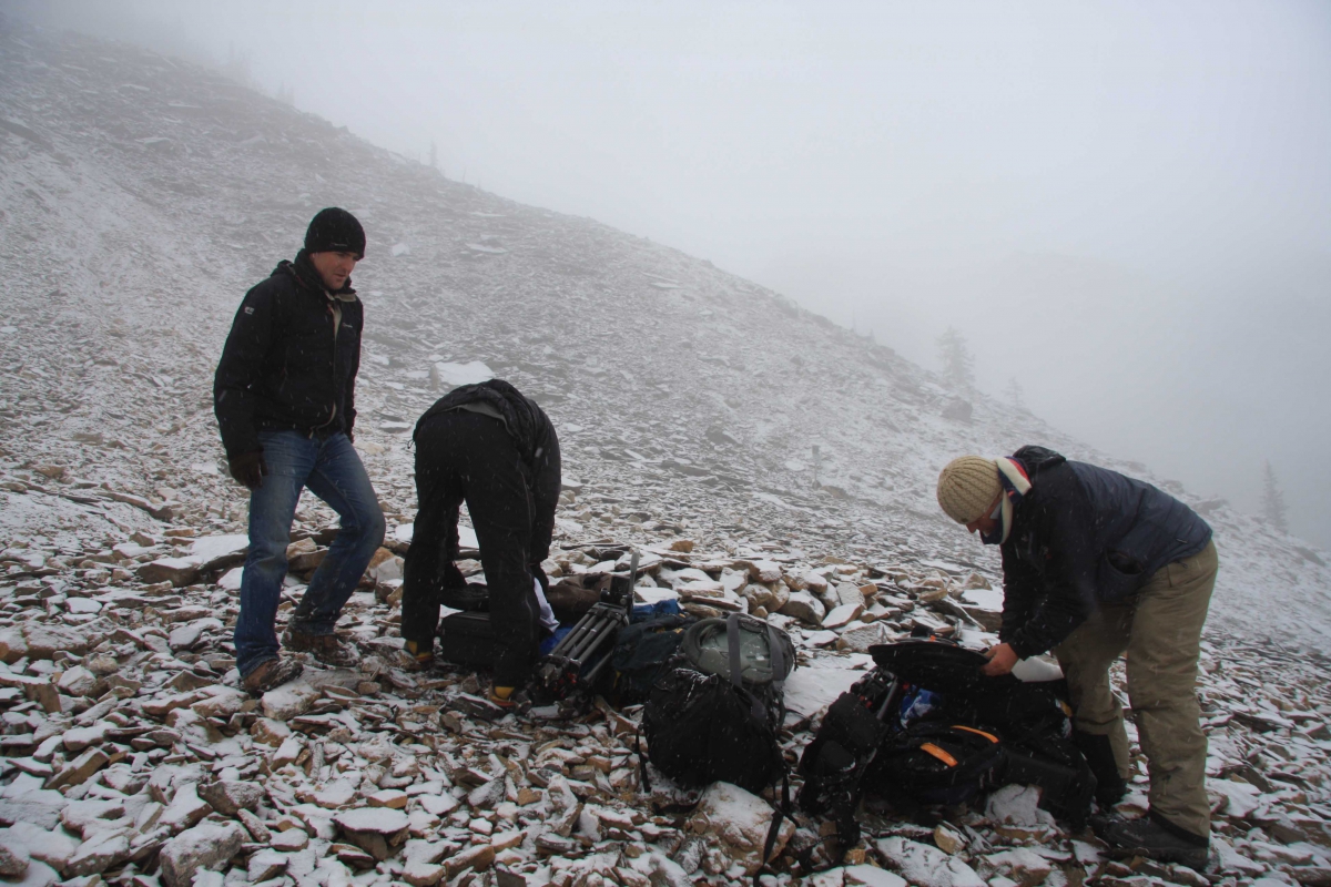 Three people in outdoor clothing standing and bending over backpacks on a rocky, snowy slope with foggy gray sky.