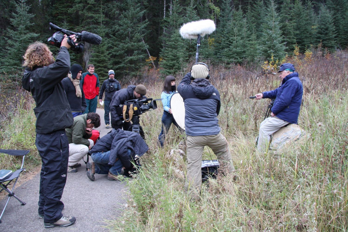 Videography crew filming a person sitting on a stool in a forest clearing surrounded by tall grass and trees.