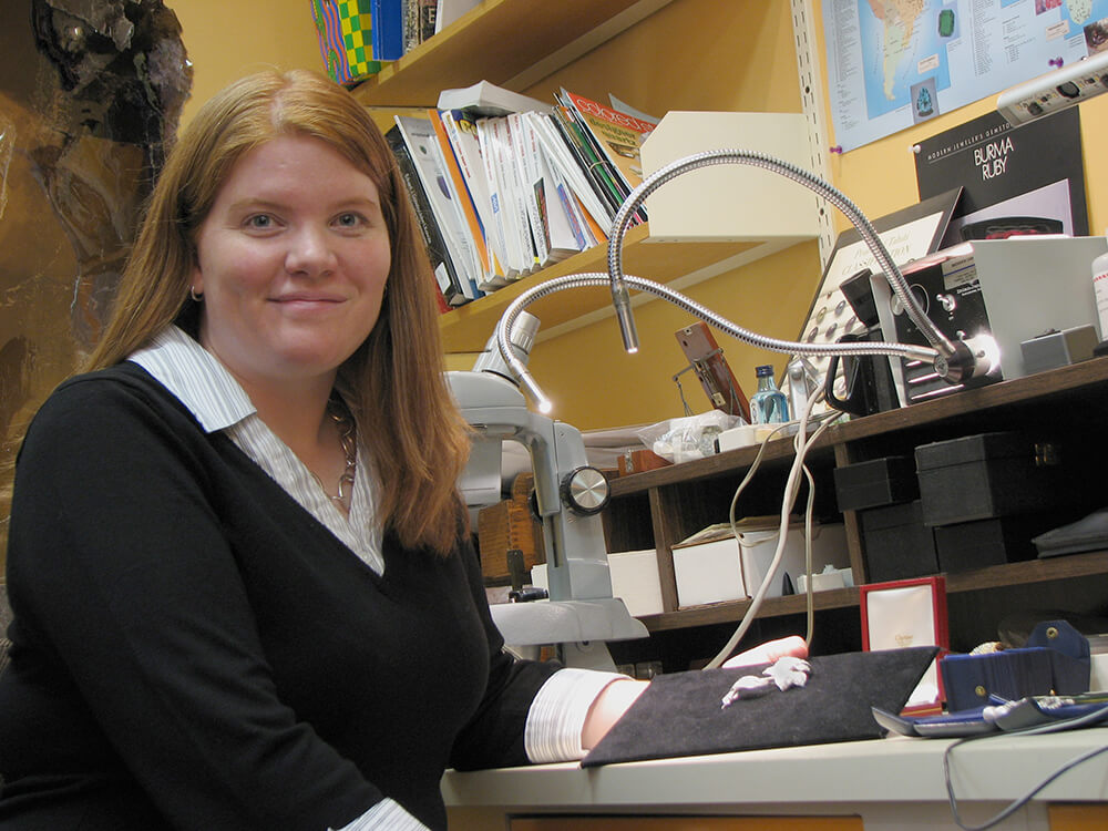 A woman with light brown hair and a black sweater sits by a laboratory setup with wires, a monitor, and printed materials.