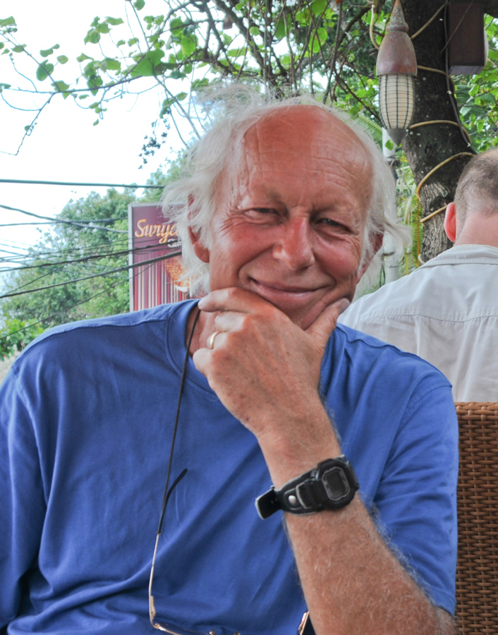 An elderly man with white hair and a beard, wearing a blue shirt and a black watch, resting his chin on his hand and smiling outdoors.