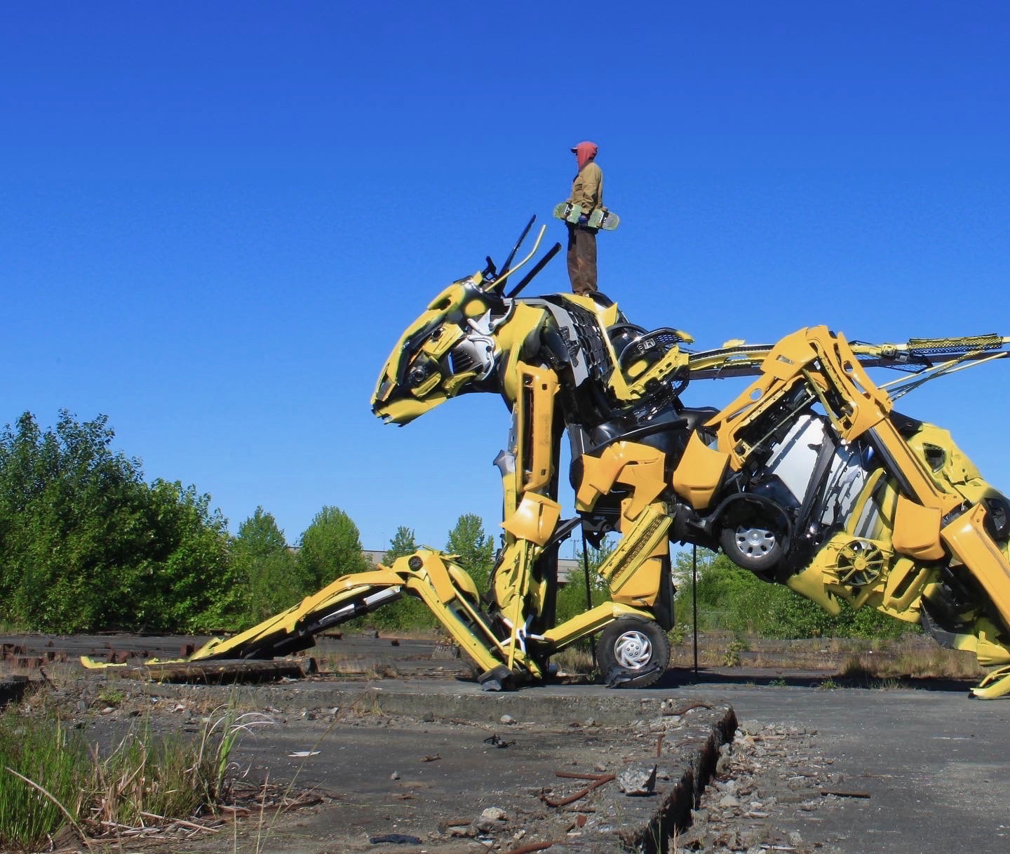 Large yellow mechanical dinosaur sculpture made from industrial parts on a wooden platform outdoors under a clear blue sky.