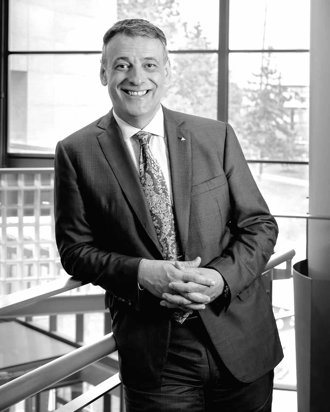 Man in a suit and patterned tie standing indoors in front of large windows with a handrail visible behind him.