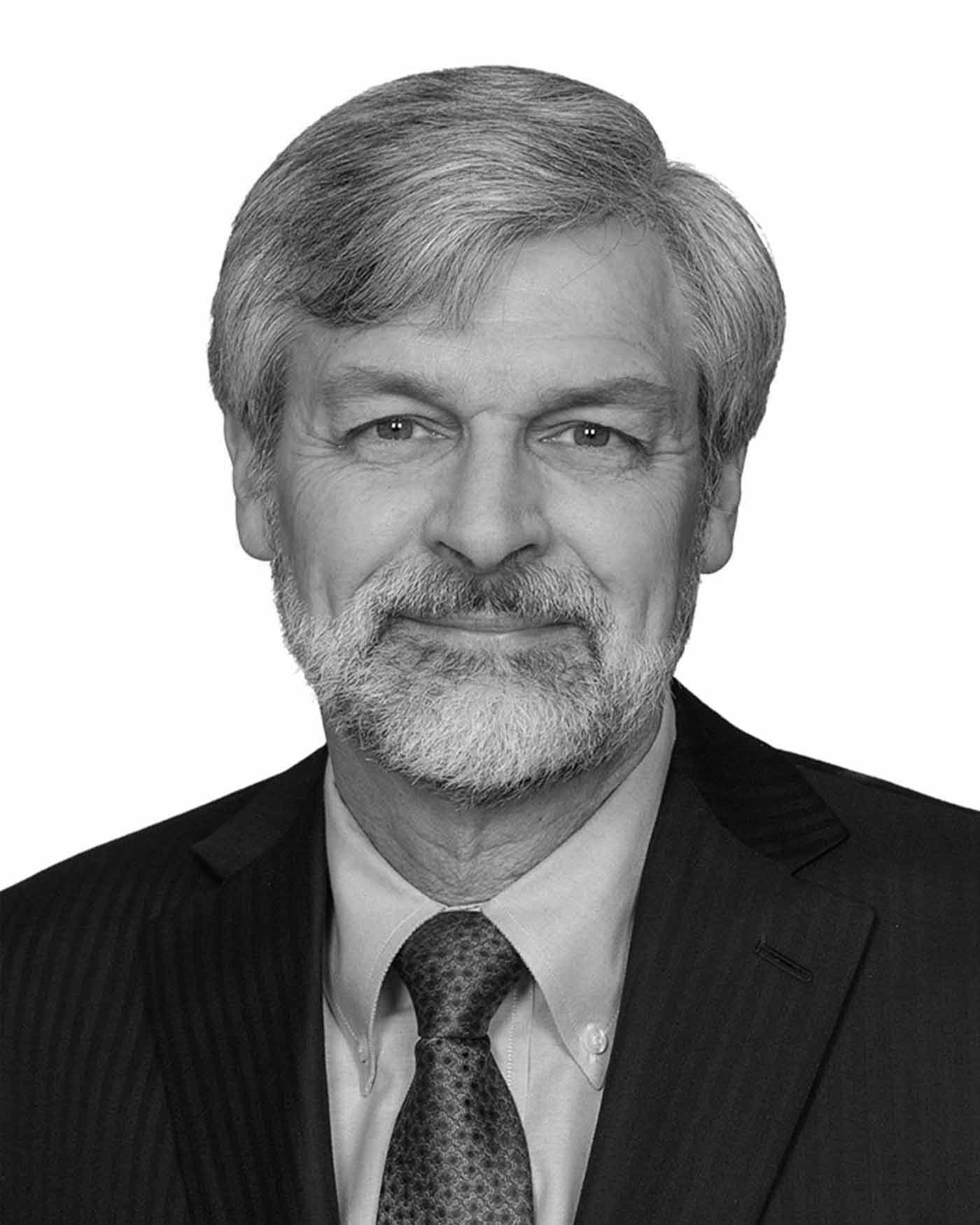 Portrait of a man with gray hair and beard, wearing a dark pinstripe suit, patterned tie, and light shirt, against a plain white background.