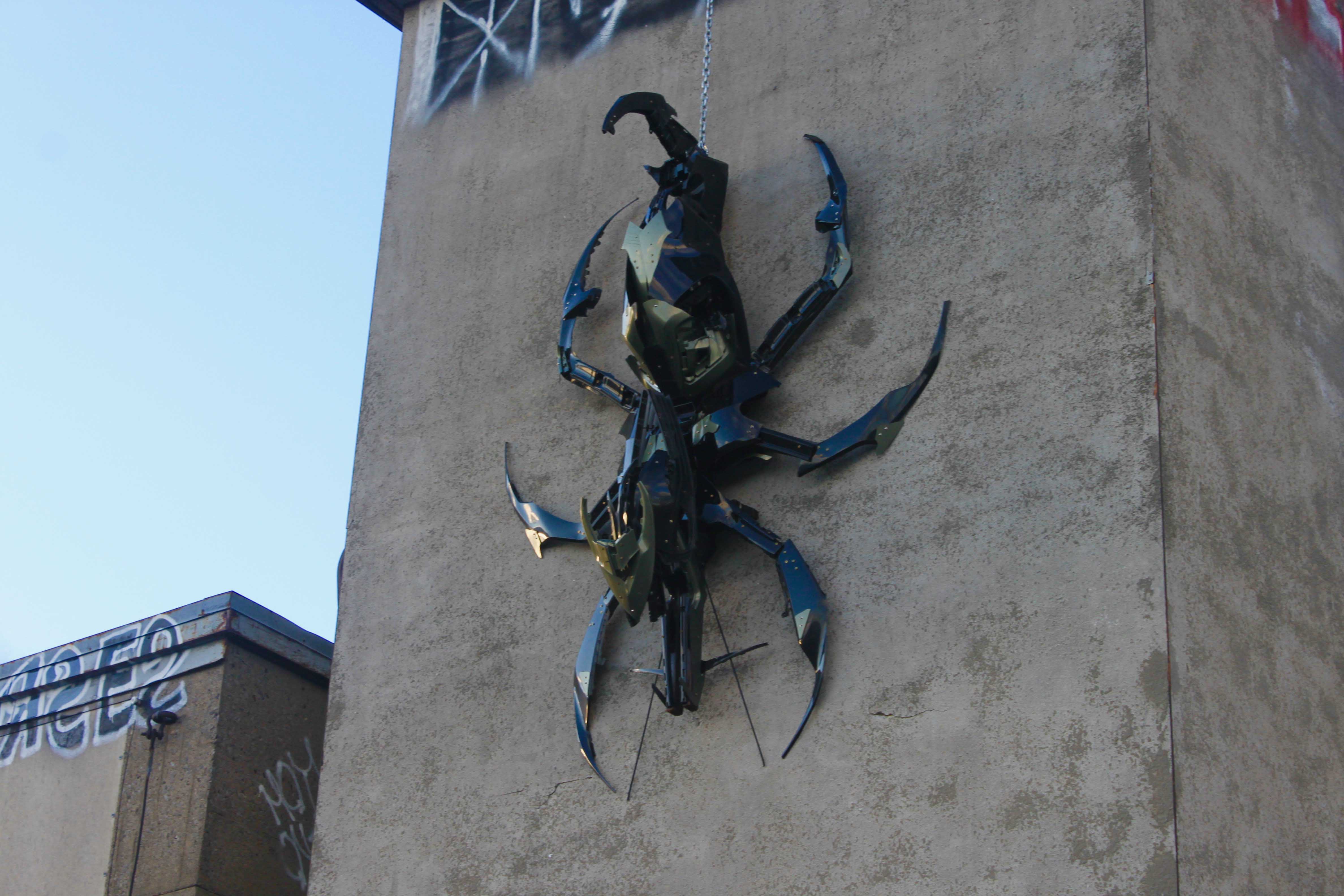 Large blue and black metal beetle sculpture mounted on a beige concrete building wall.