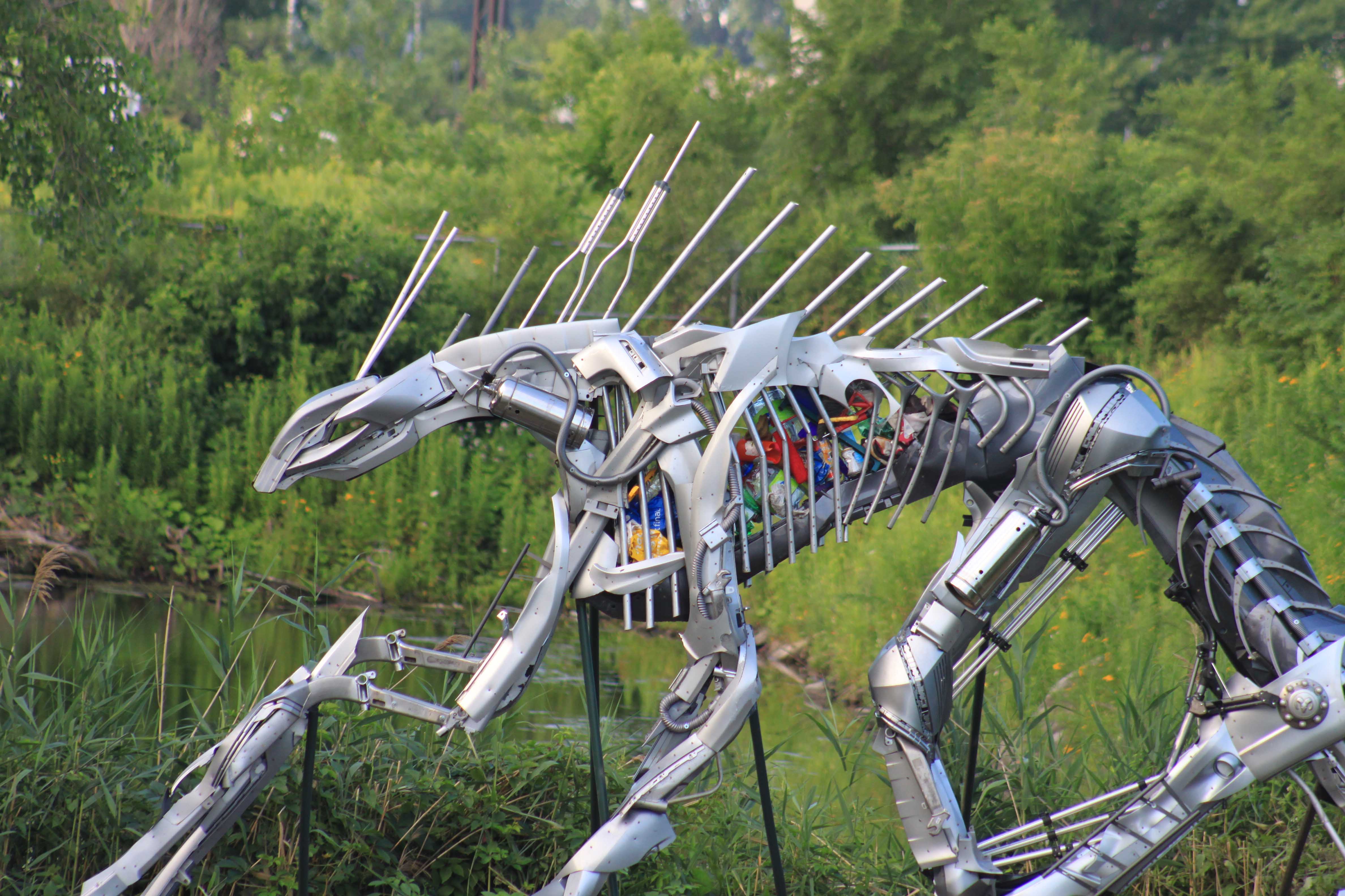 Large metallic sculpture of an insect with multiple legs and antennae, set outdoors with greenery in the background.