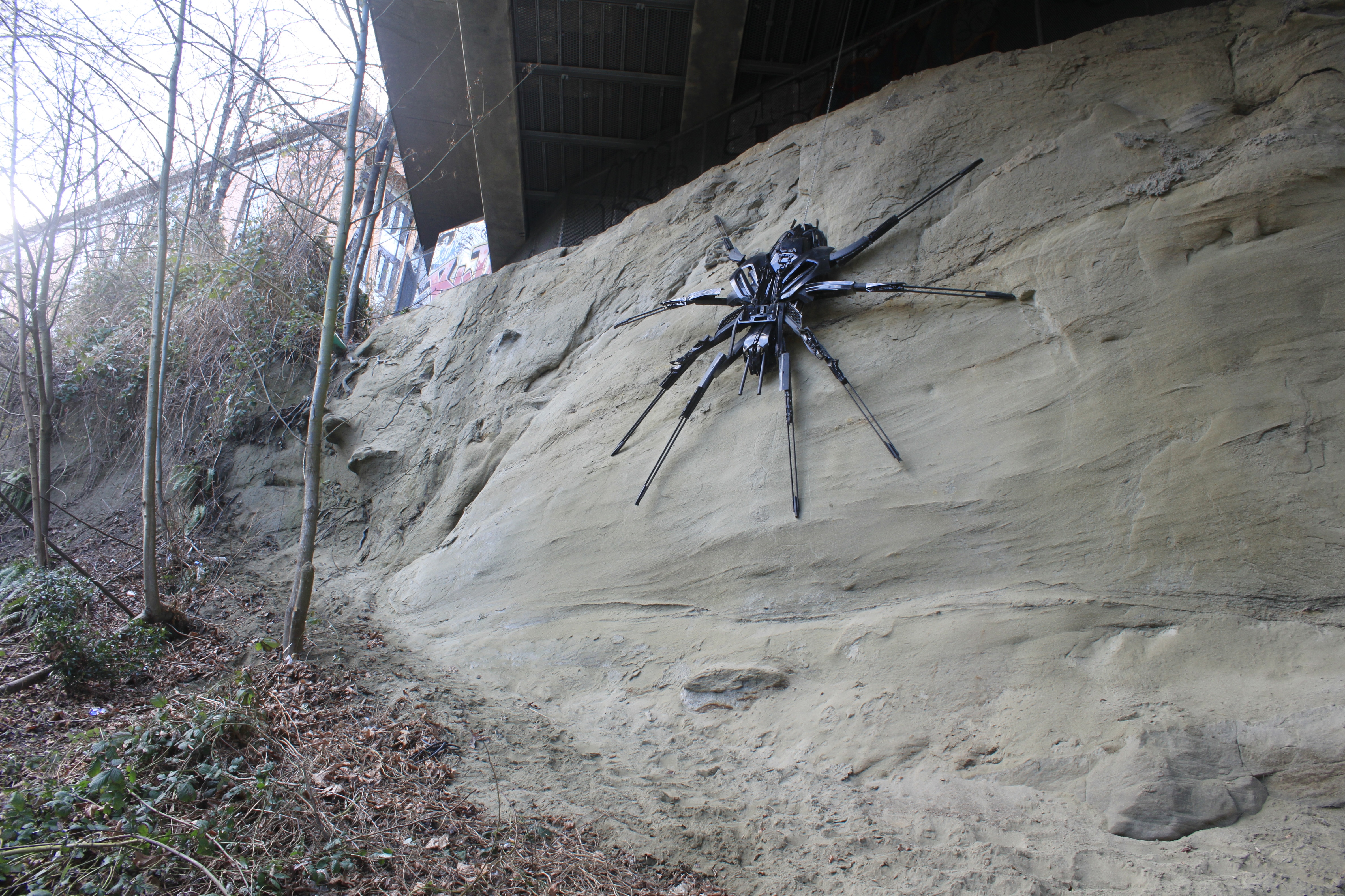 Large black spider sculpture mounted on a sandy rock cliff outdoors among bare trees.