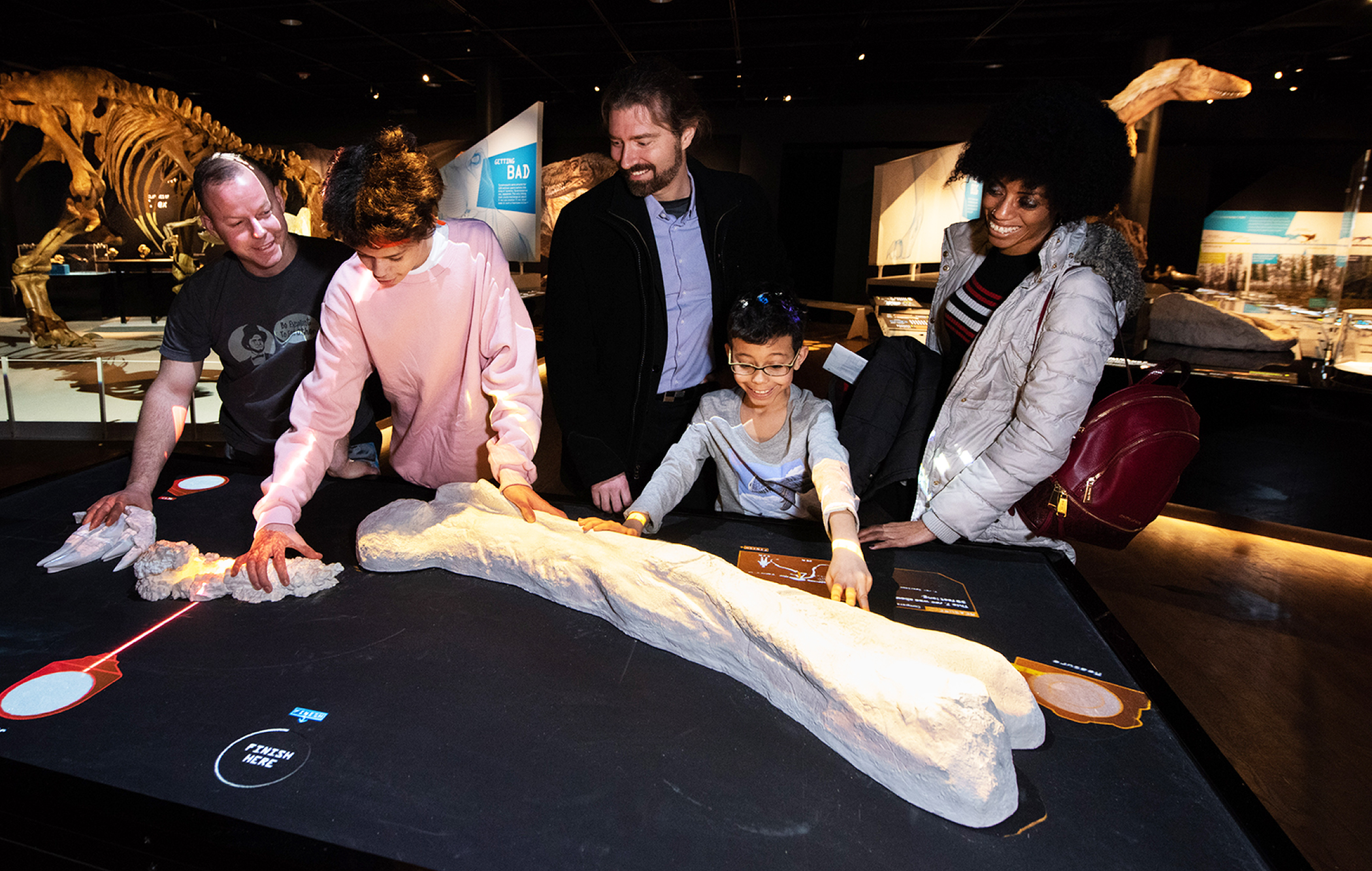 Five people, including a child, examine a large dinosaur bone displayed on a black table in a dimly lit museum gallery.