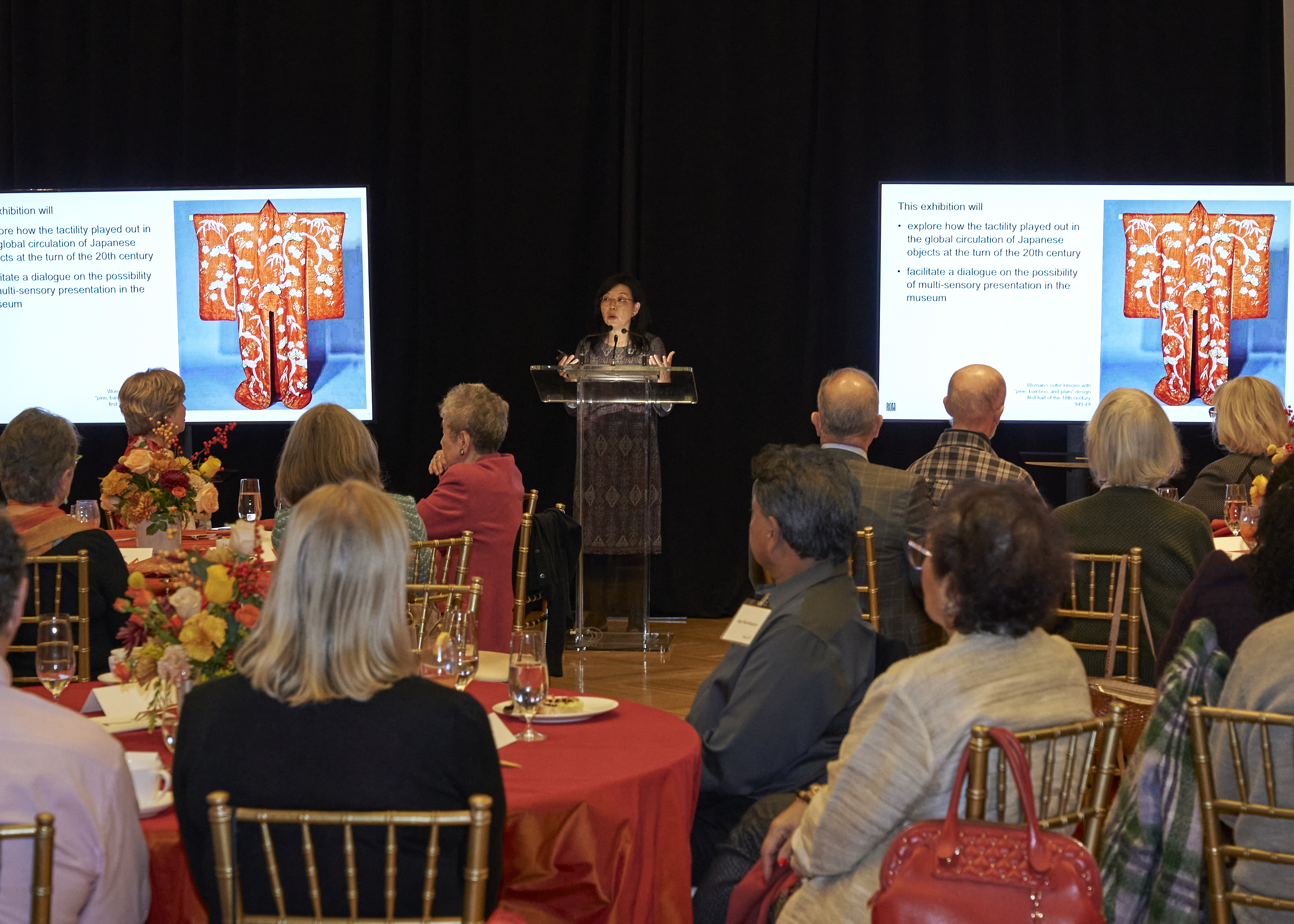 A speaker at a podium presents to an audience seated at round tables with two large screens displaying an artifact and text.