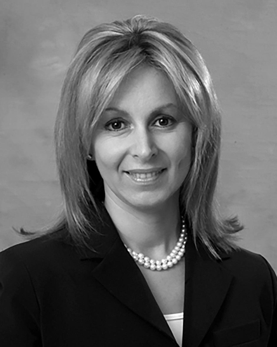 Portrait of a woman with shoulder-length blonde hair, wearing a dark blazer and a pearl necklace, on a plain background.