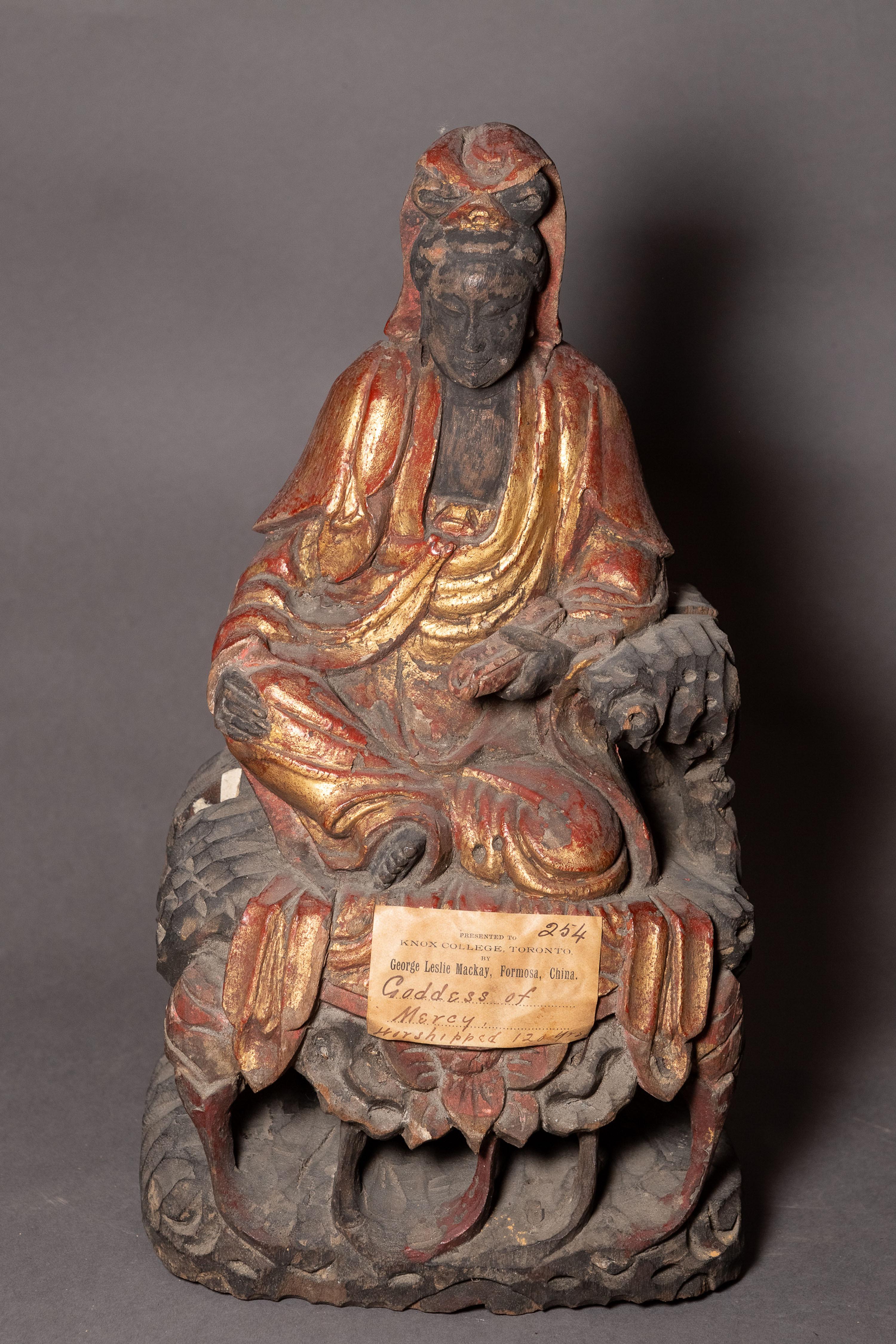 Seated figure statue with a beard, wearing a robe and holding an object, with a worn label on the base.