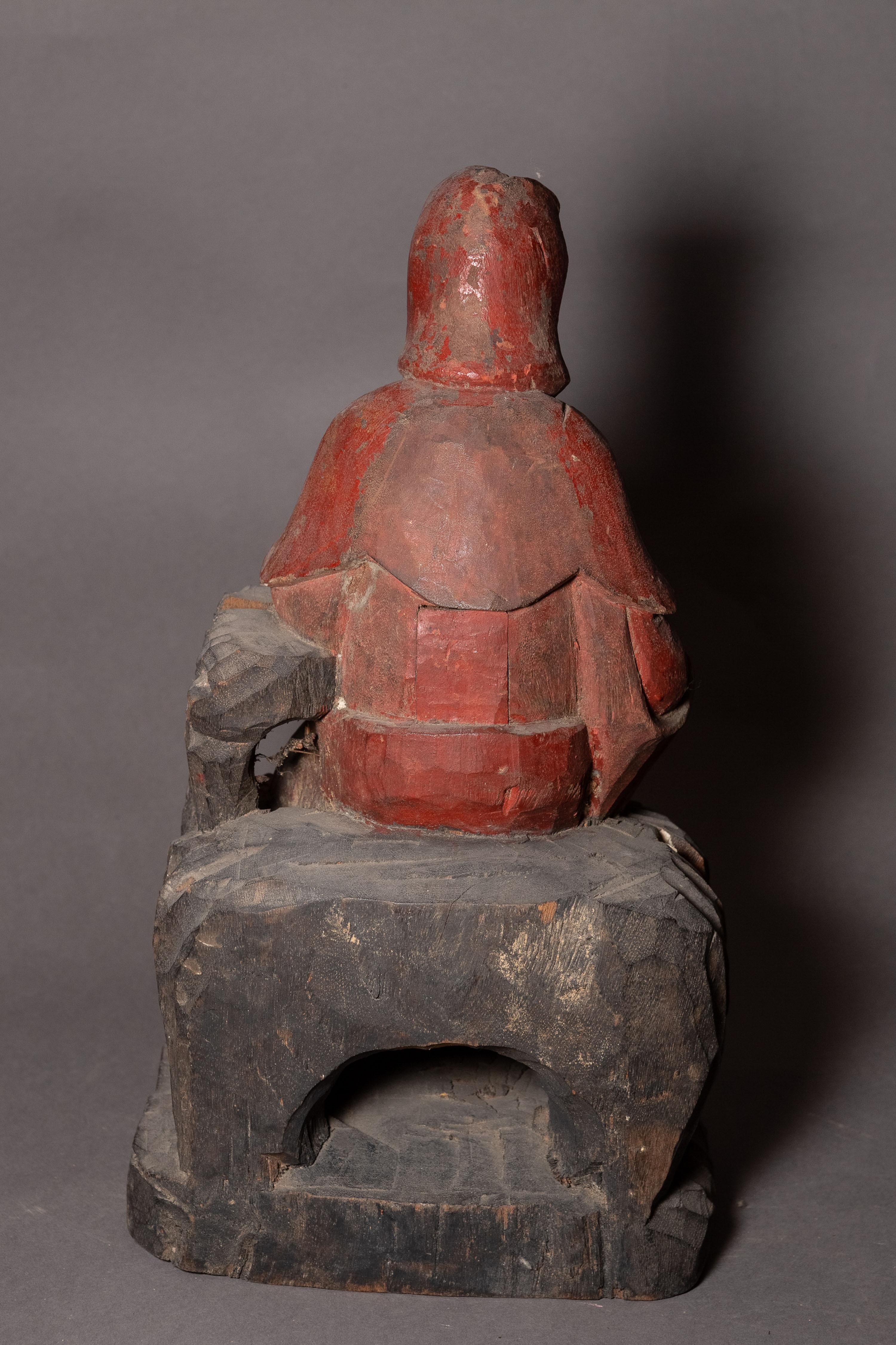 Wooden sculpture painted red and black depicting a seated figure with a rounded back and covered head.