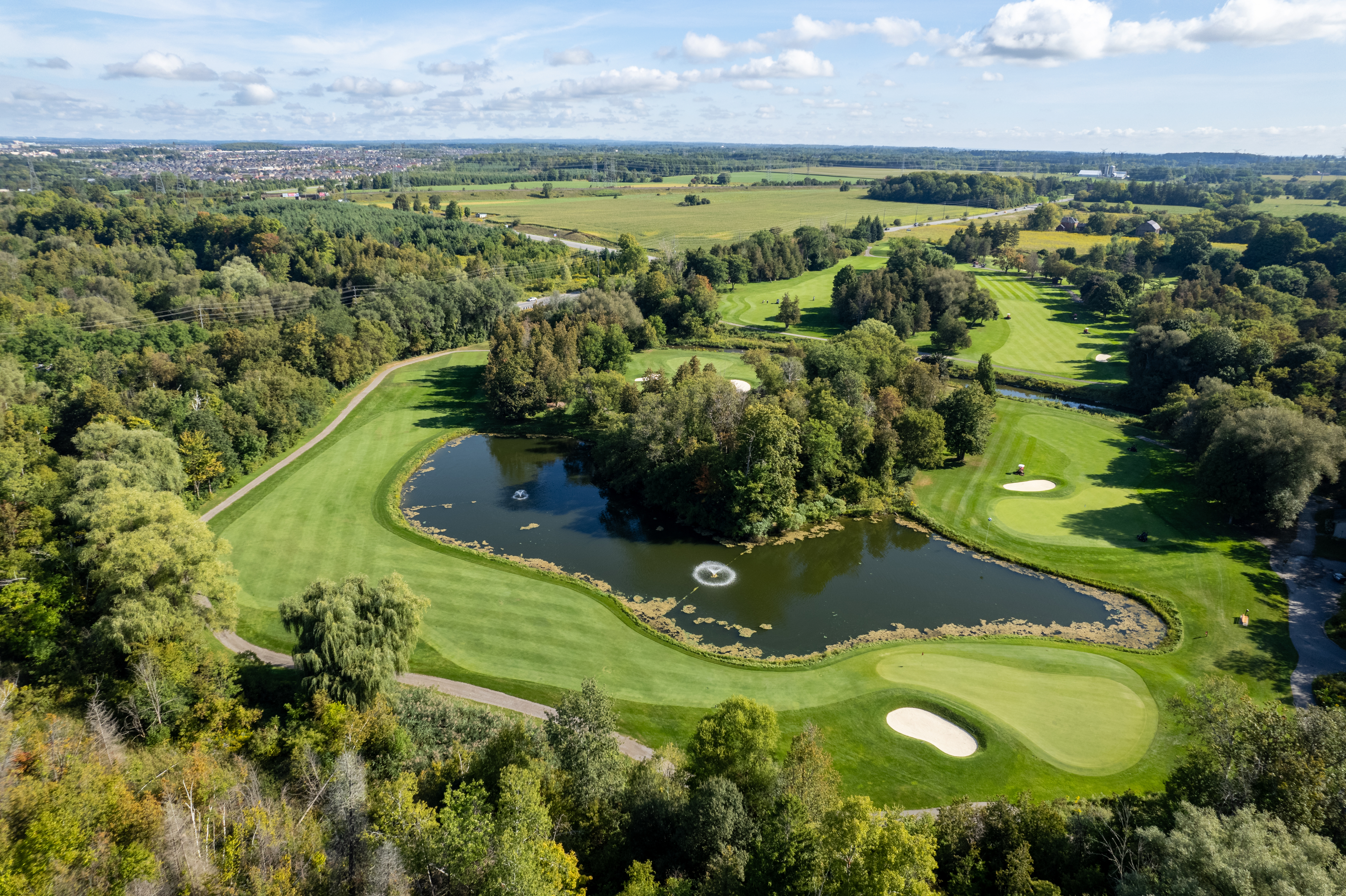 Aerial view of a golf course with a water hazard, sand traps, and surrounding trees under a blue sky.