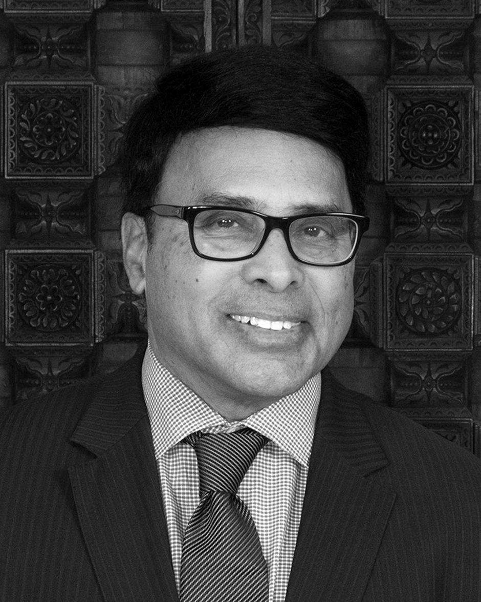 No visible text; black-and-white portrait of a man wearing glasses, a suit jacket, a patterned tie, and smiling.