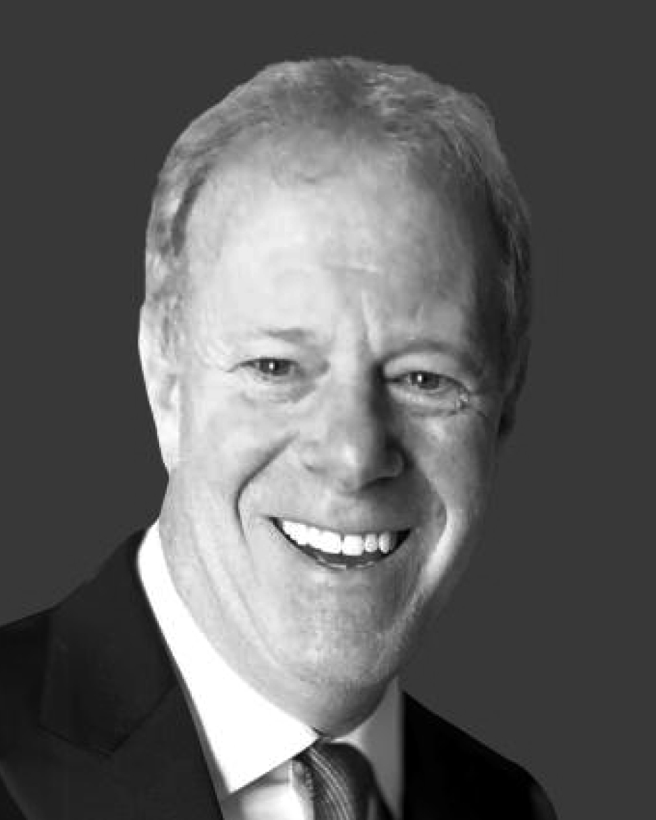 Portrait of an older man with short grey hair, smiling, wearing a dark suit and white shirt, on a grey background.
