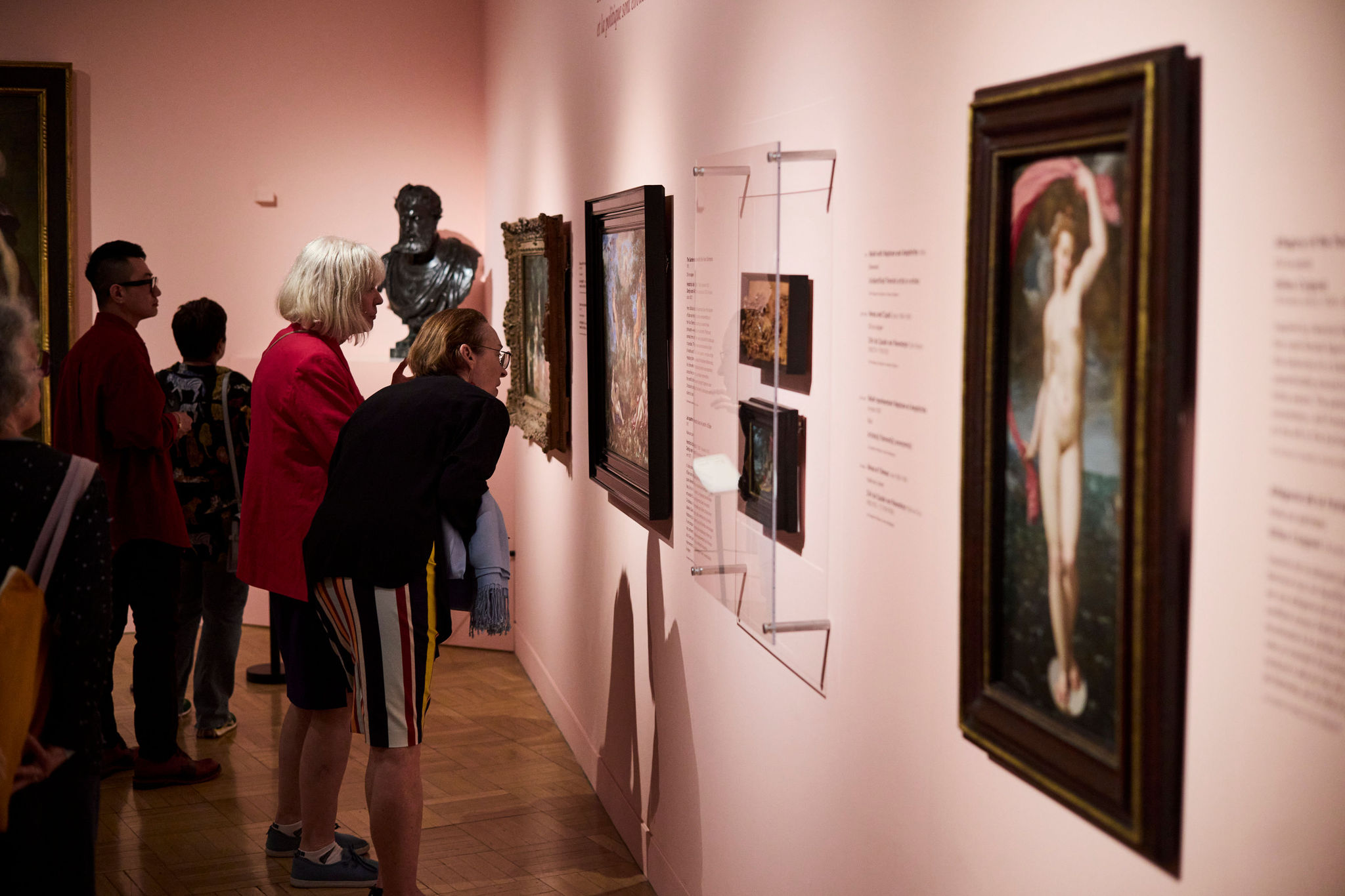Several people closely observing framed artworks displayed on a museum wall with soft pink lighting.