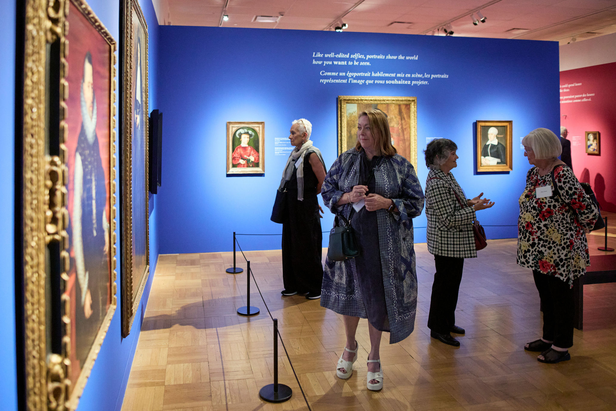 Five adults stand or walk inside an art gallery with blue and pink walls displaying framed paintings.