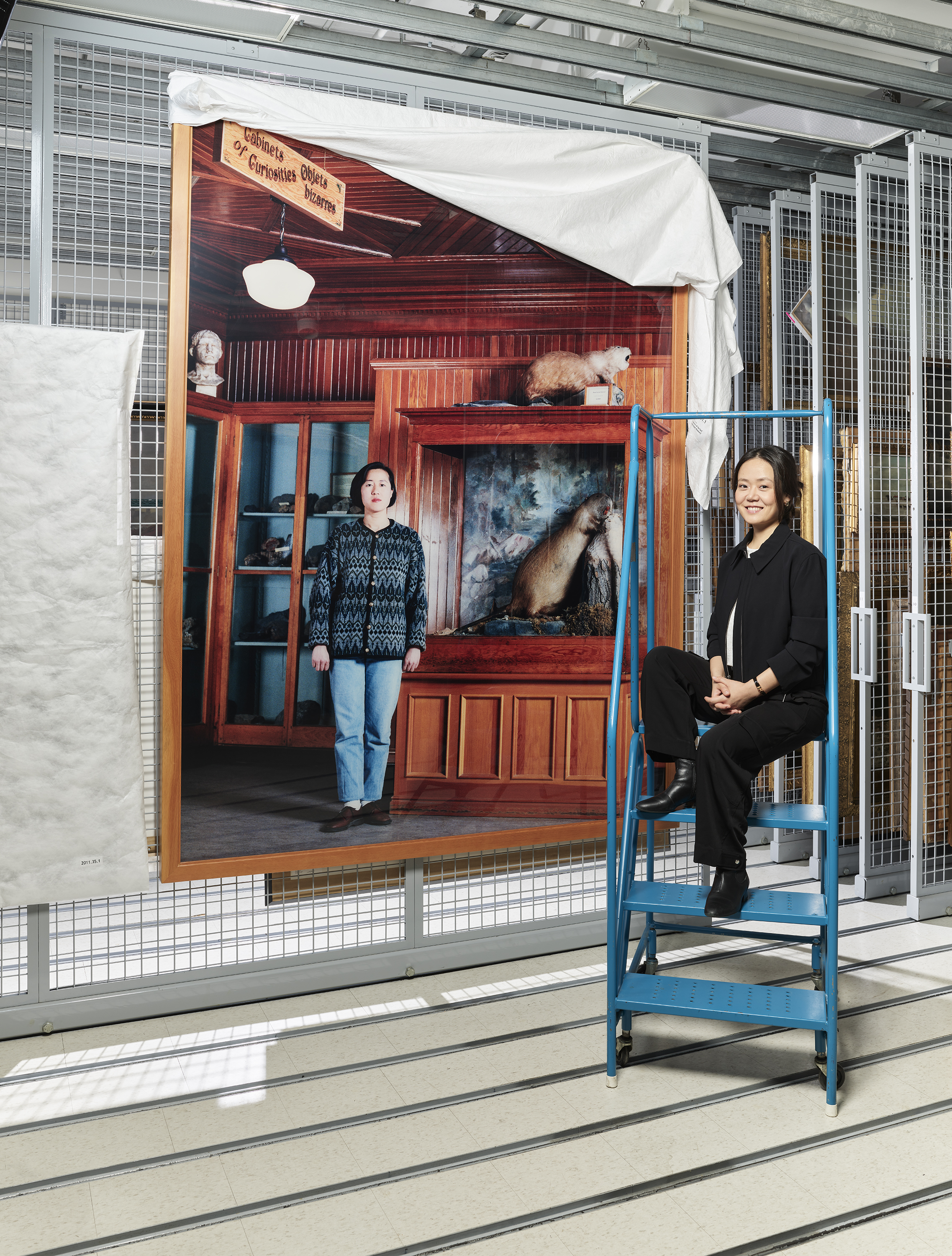 Two women pose near a large photographic print hanging from a metal grid; one woman stands in front while the other sits on a ladder.