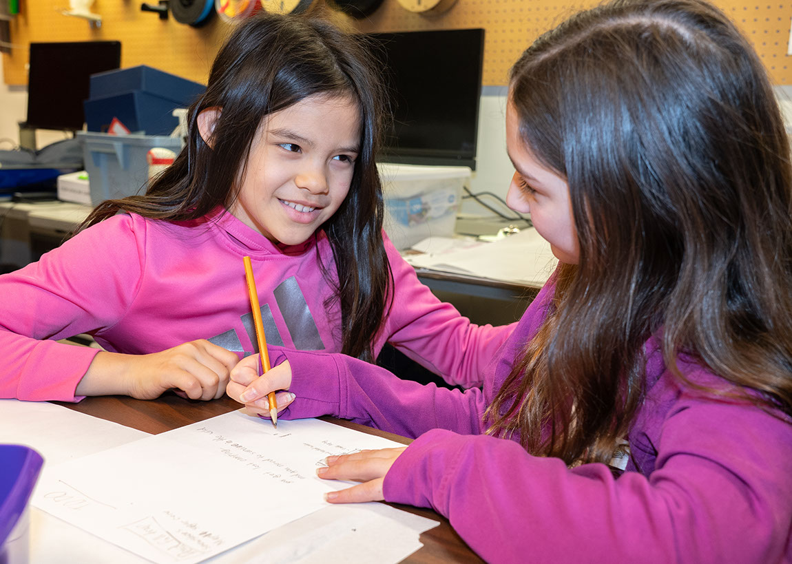Two smiling young students look at each other. One is holding a pencil and writing.