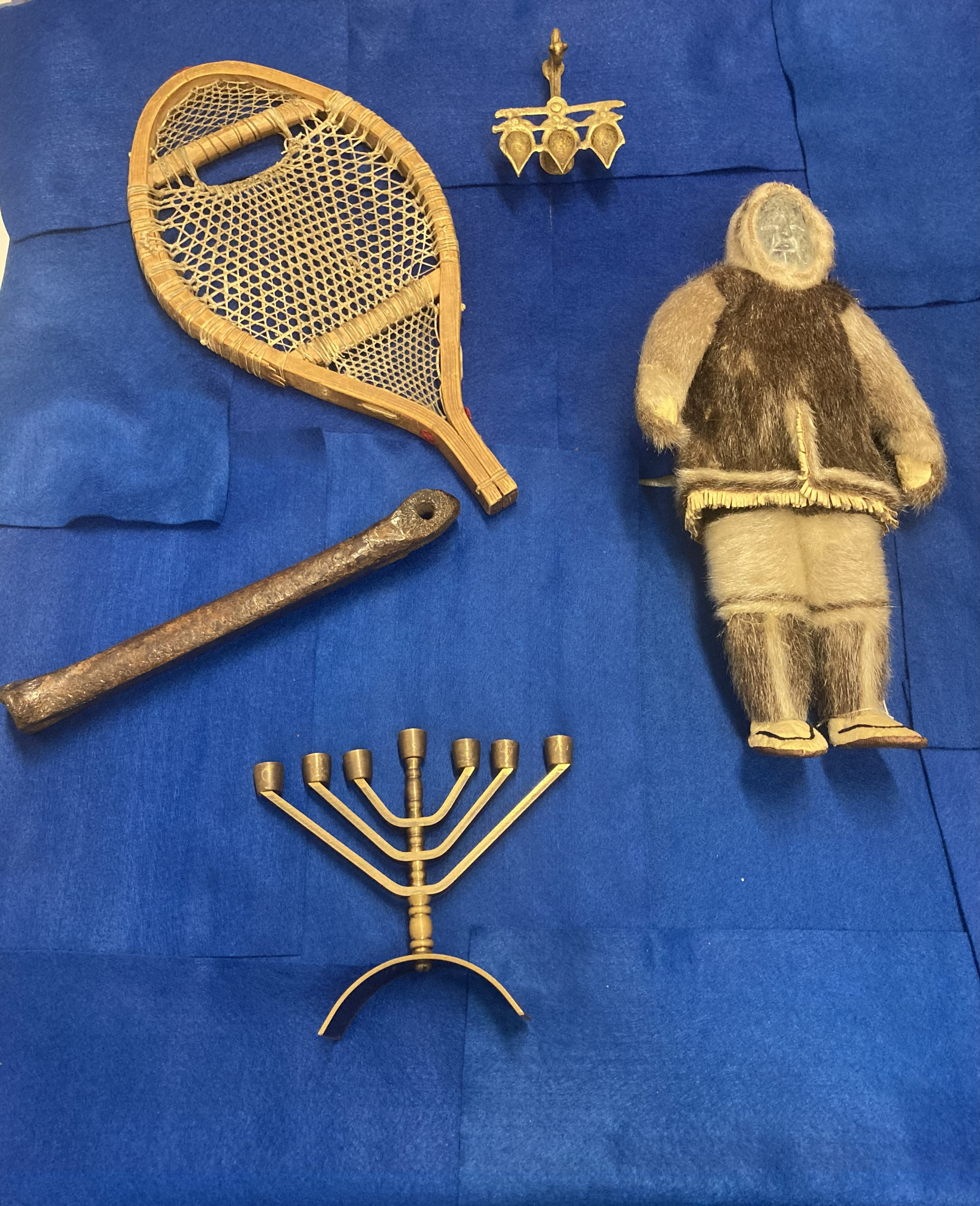 Sélection d'objets sur fond bleu.  Les objets comprennent une poupée inuit en pierre et en peau de phoque, une Hannukiah, un patin médiéval en os, une raquette et une lampe diya.
