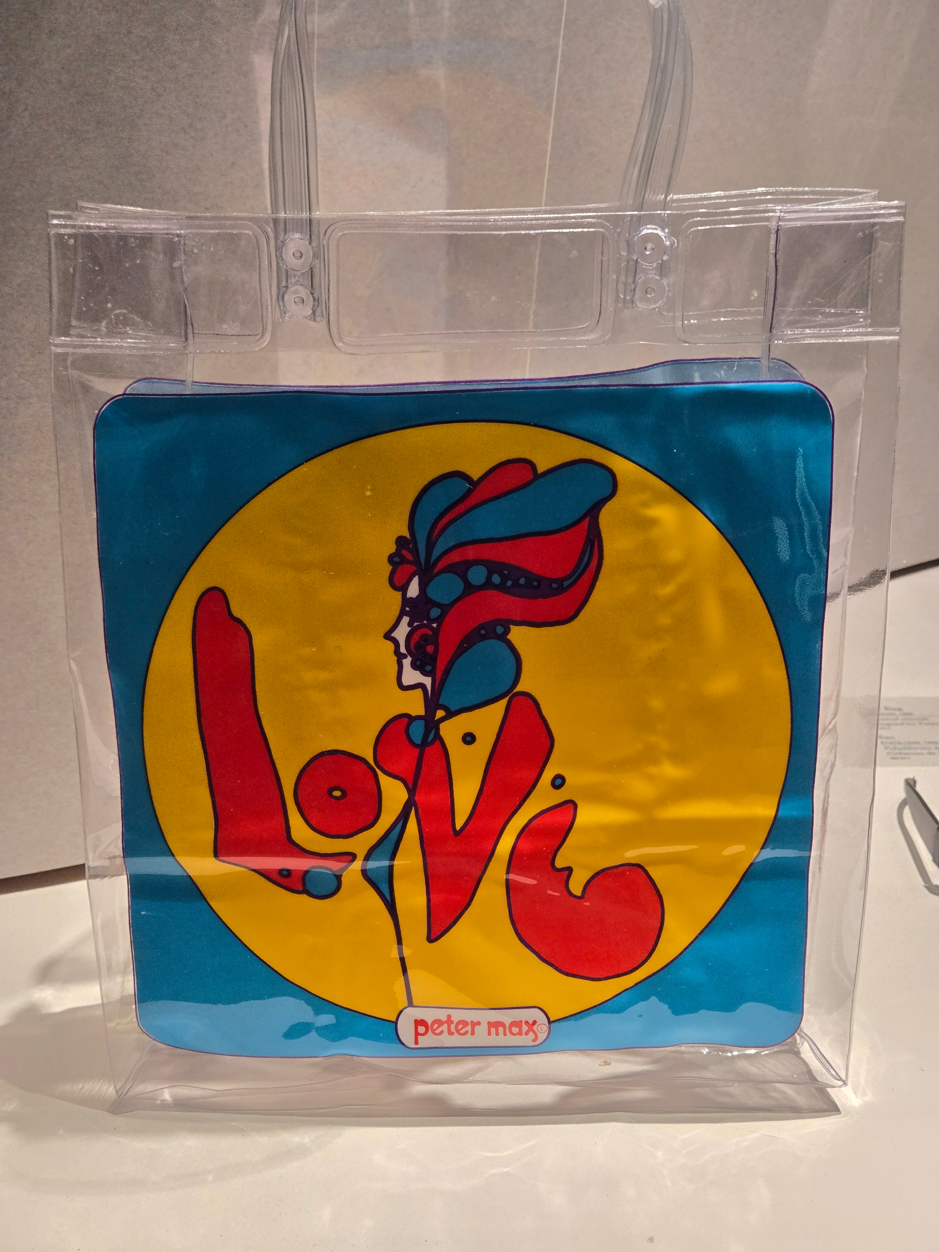 Un sac en plastique transparent dans un coffret de musée. Sur le devant du sac figure l'image d'une femme dans un cercle jaune sur fond bleu. Les armes et le manteau de la femme épellent également le mot AMOUR. Peter max est imprimé au bas du sac.