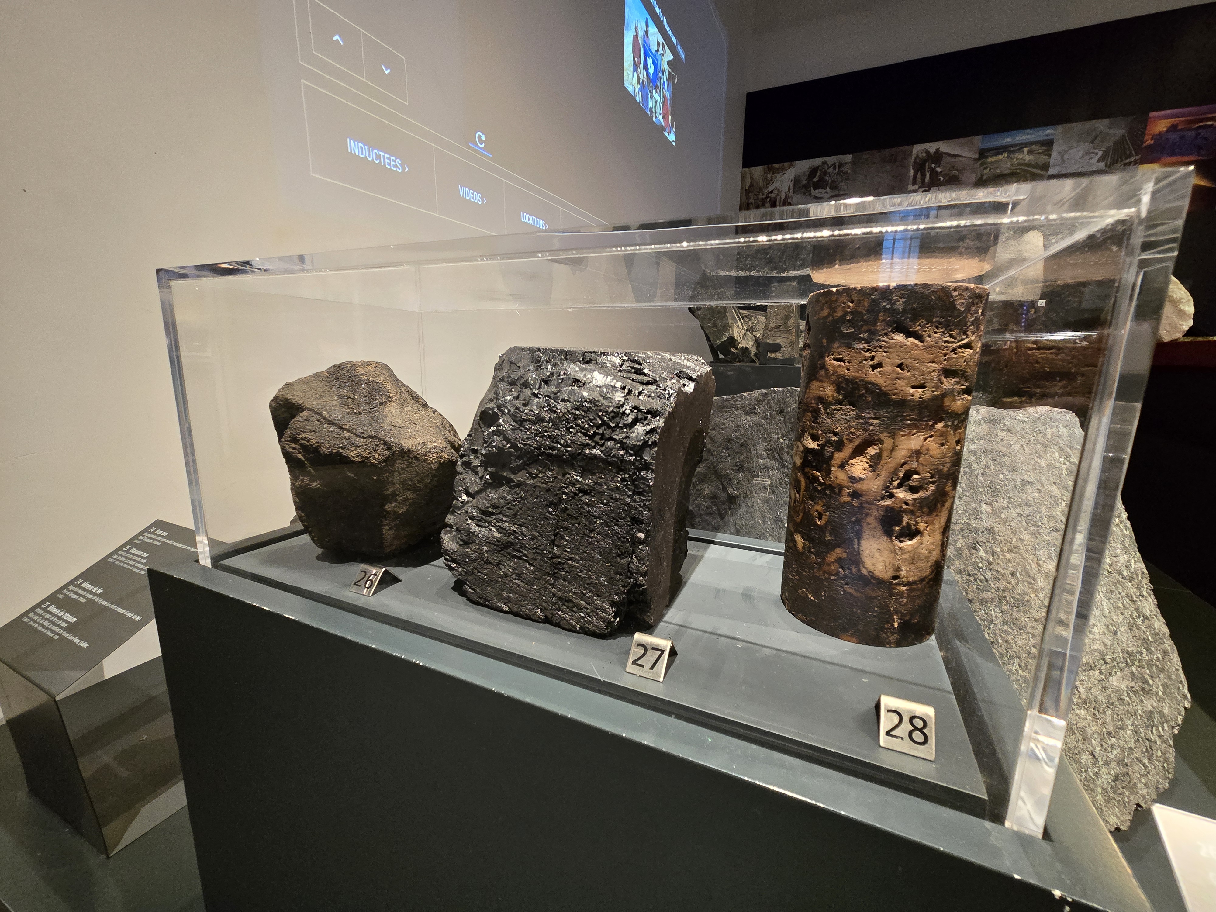 Dans la galerie d'un musée, une vitrine contient trois roches. L'une est brune/noire terne et arrondie, l'autre est noire et brillante, et la troisième présente des tons bruns avec des taches plus claires sur la roche plus foncée.