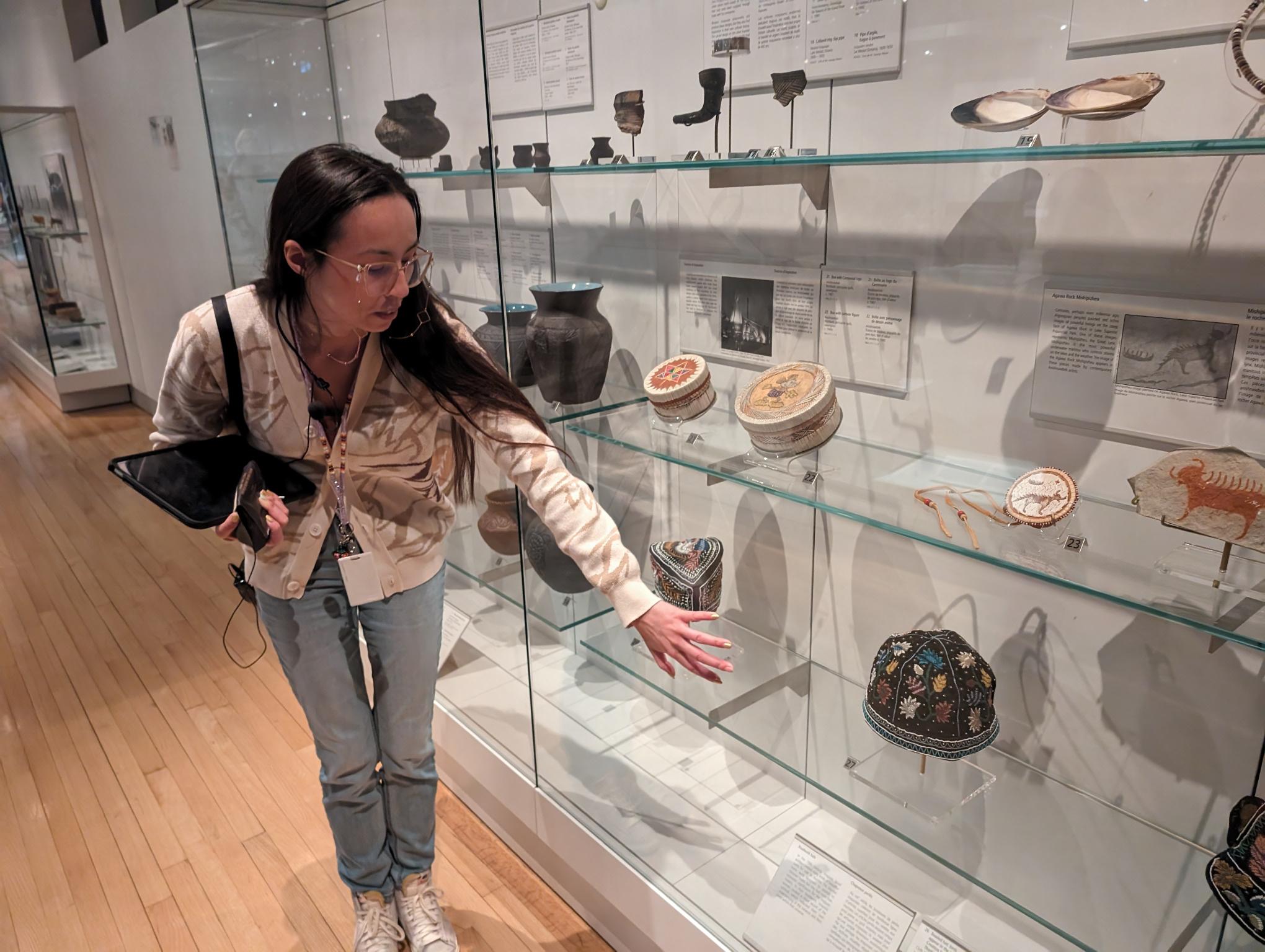 An Indigenous woman with long, dark hair and glasses stands by a museum case filled with belongings showing beadwork, quillwork, and other artistic traditions.