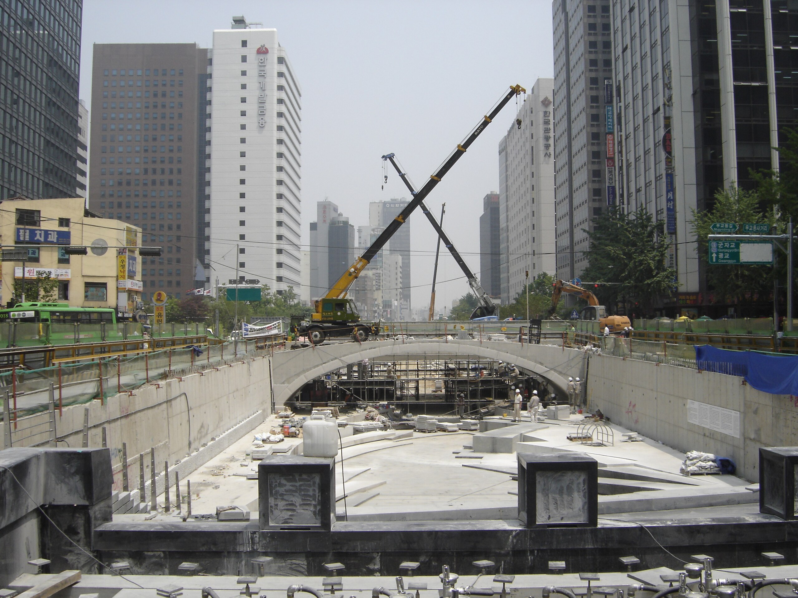 Construction site of an underground structure between tall buildings with cranes and rebar visible.