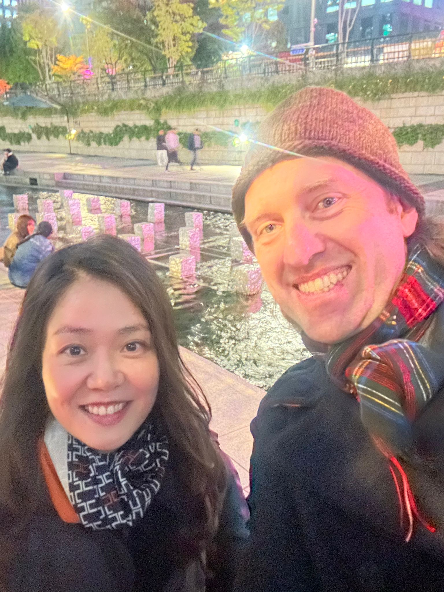 Two people smiling outdoors at night with festive string lights and reflections on water behind them.