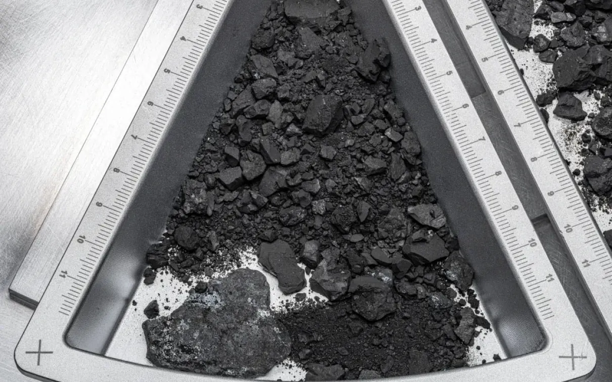 Pile of black coal-like chunks inside a white triangular container marked with a "+" sign.