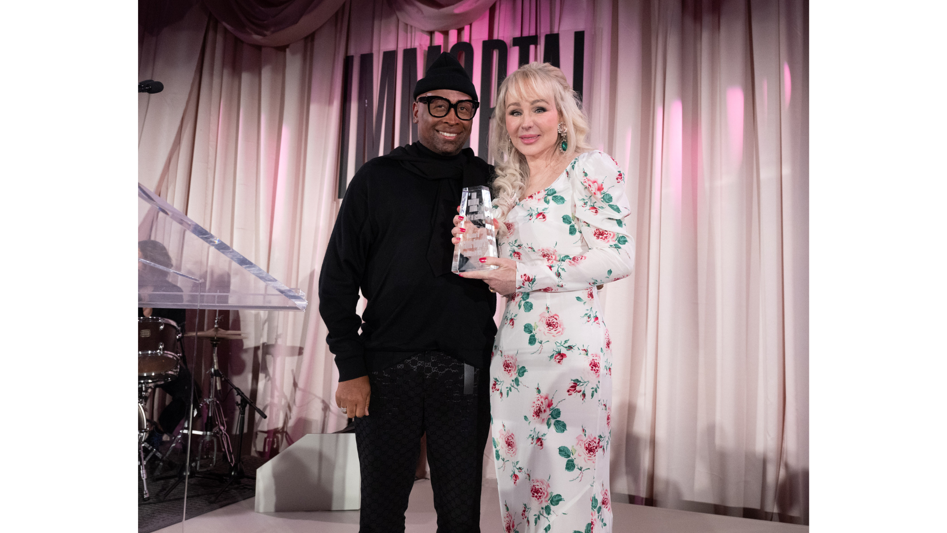 A man in a black outfit and a woman in a white floral dress stand closely together on a stage with a curtain backdrop.