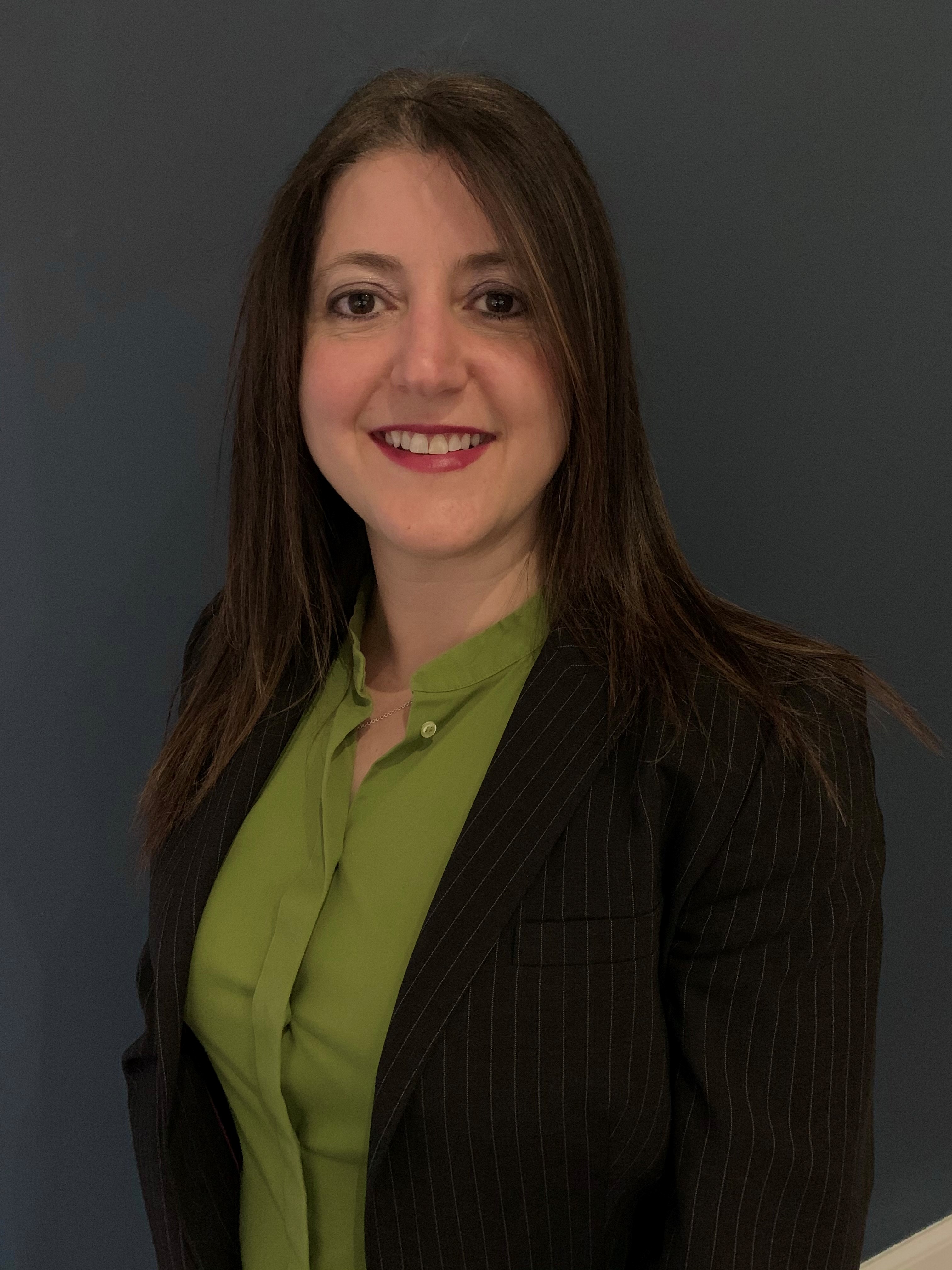 Portrait of a woman with long brown hair, wearing a black pinstripe blazer over a green blouse, against a dark background.