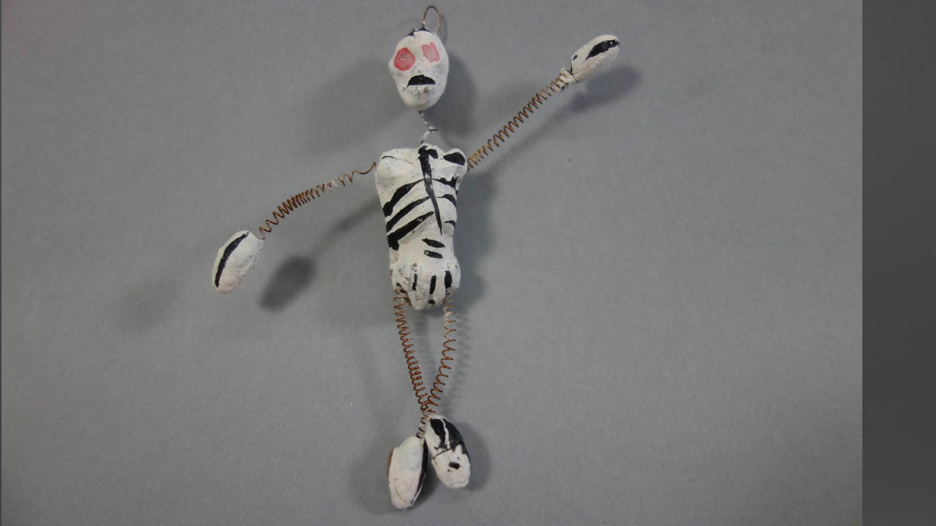Small handcrafted figure with a white body painted with black markings, red mouth, and orange eyes, posed against a gray background.