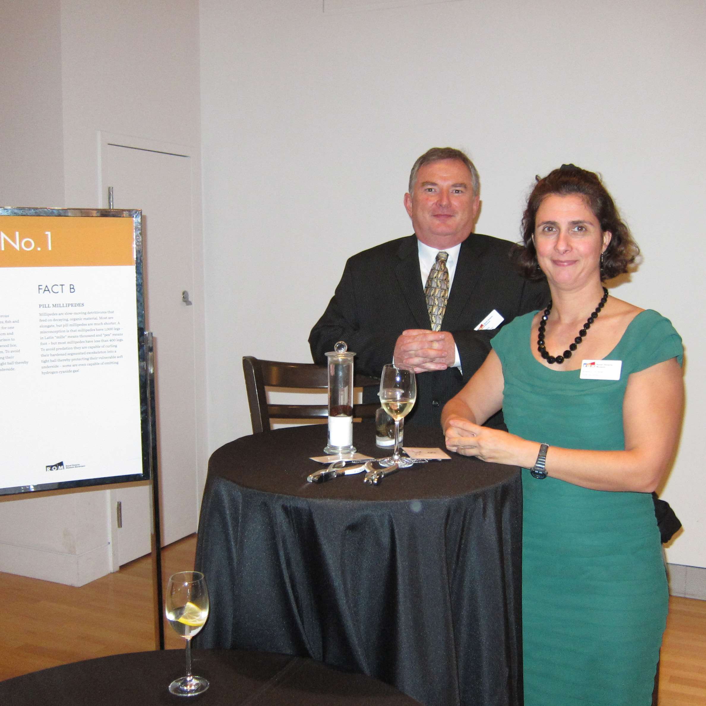 Two people stand next to a high round table with glasses and drinks in a room with a white wall and a sign labeled "No. 1."