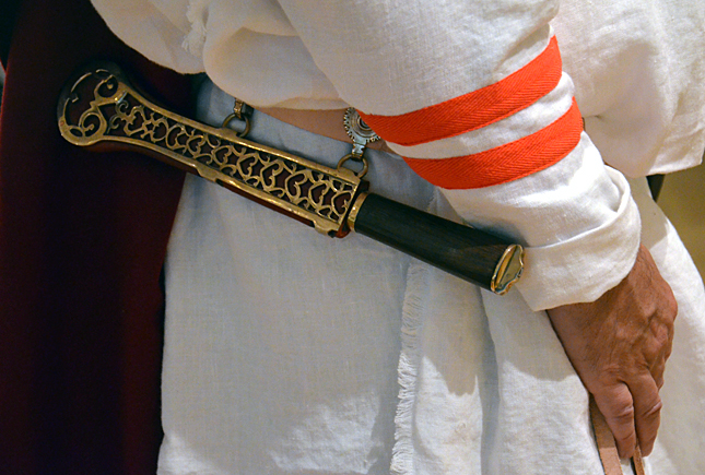Close-up of a person wearing a white garment with red stripes on the sleeve and a decorative sheath with gold patterns holding a wooden stick.