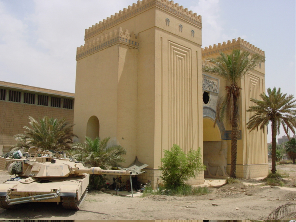 Two military tanks parked near a large beige building with decorative square patterns and palm trees nearby.
