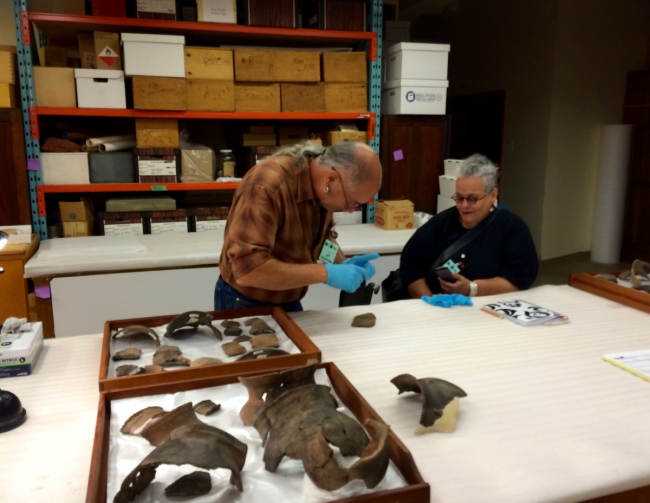 Two men examine fossilized bones spread out on a large table in a research or archival room.