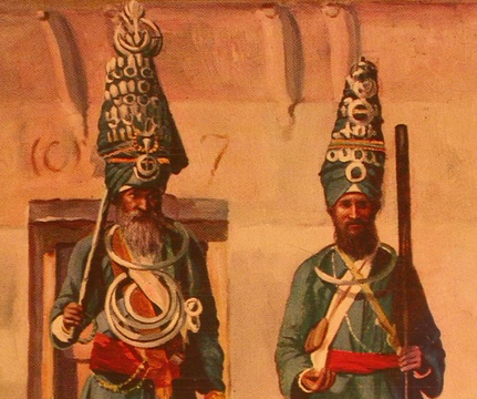 Two men in elaborate ceremonial attire with tall headdresses and intricate decorations stand against an adobe wall.