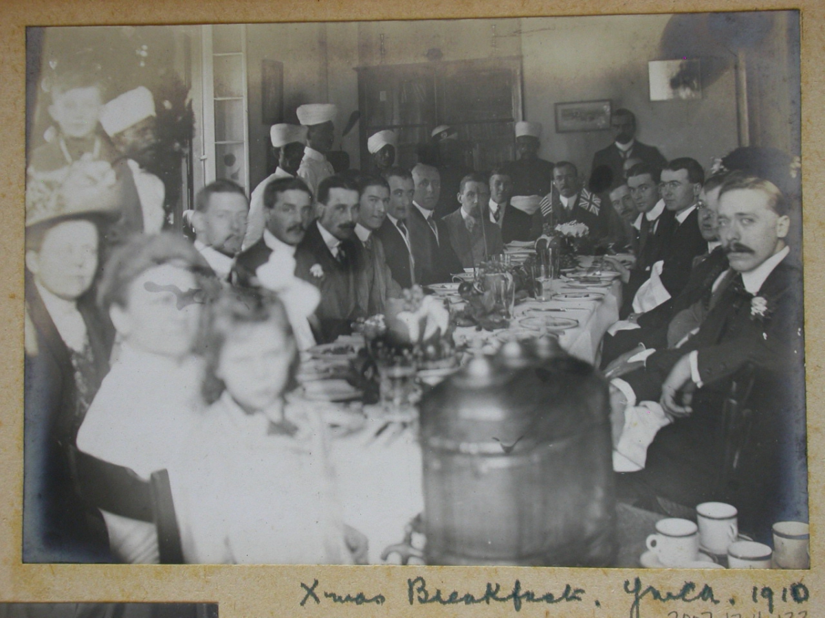 Xmas Breakfast, 1910 handwritten on the bottom border beneath a black and white photo of people seated around a long table.