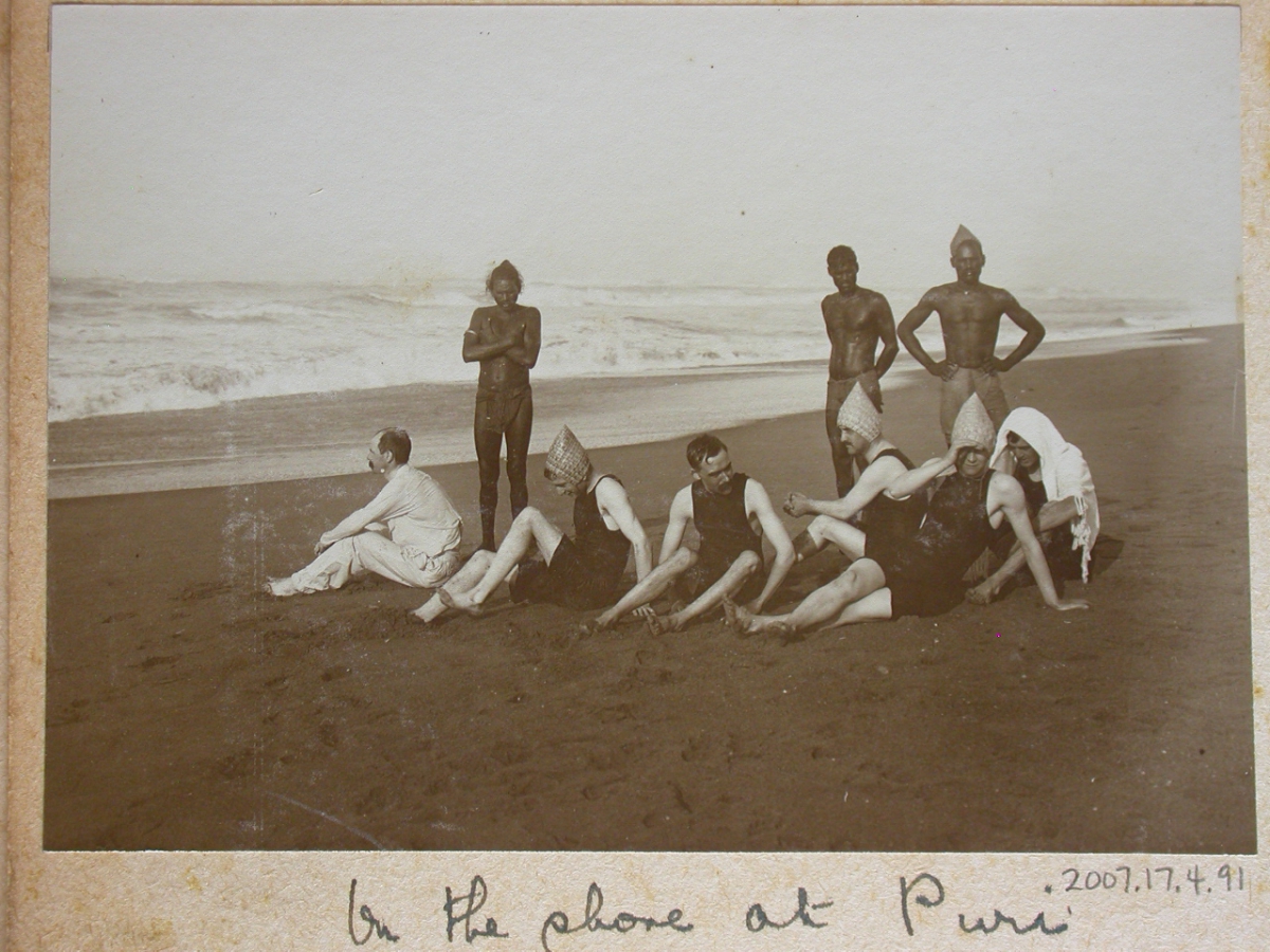 No visible text in this black-and-white photo showing eight people posing on a sandy beach with ocean waves in the background.