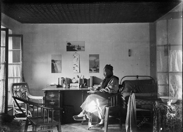 A person sitting on a bed reading a book in a sparsely furnished room with a desk, chair, and pictures on the wall.