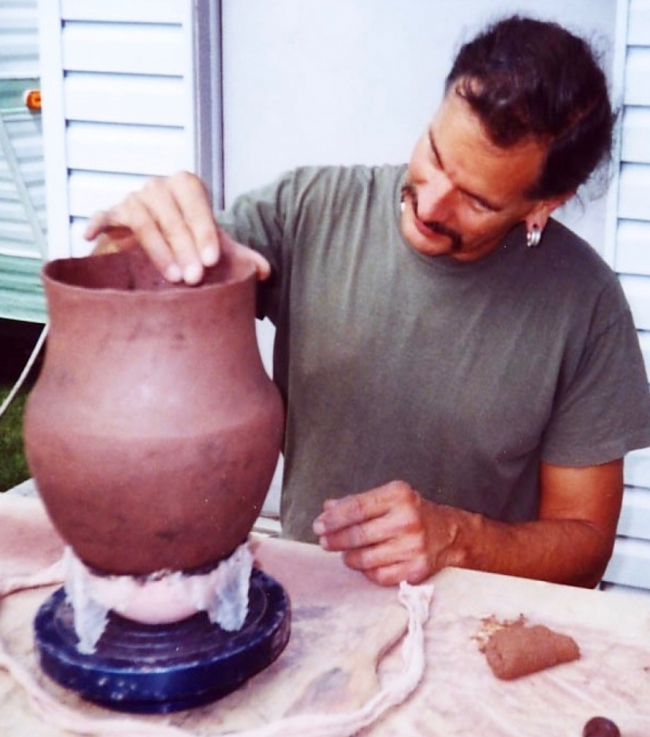 Man shaping the rim of a large brown clay pot on a pottery wheel.