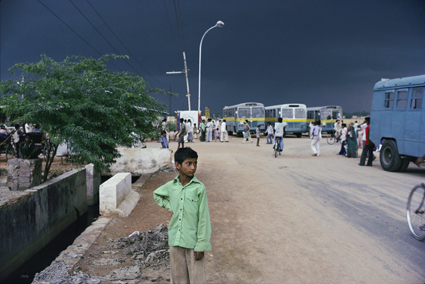 A young boy in a green shirt stands on a dirt road near buses and vehicles under a dark, cloudy sky.